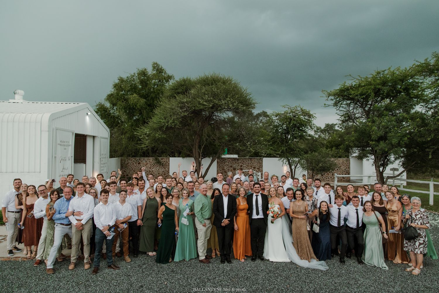Large wedding group photo outdoors with bride and groom surrounded by guests, green trees and white barn in background.