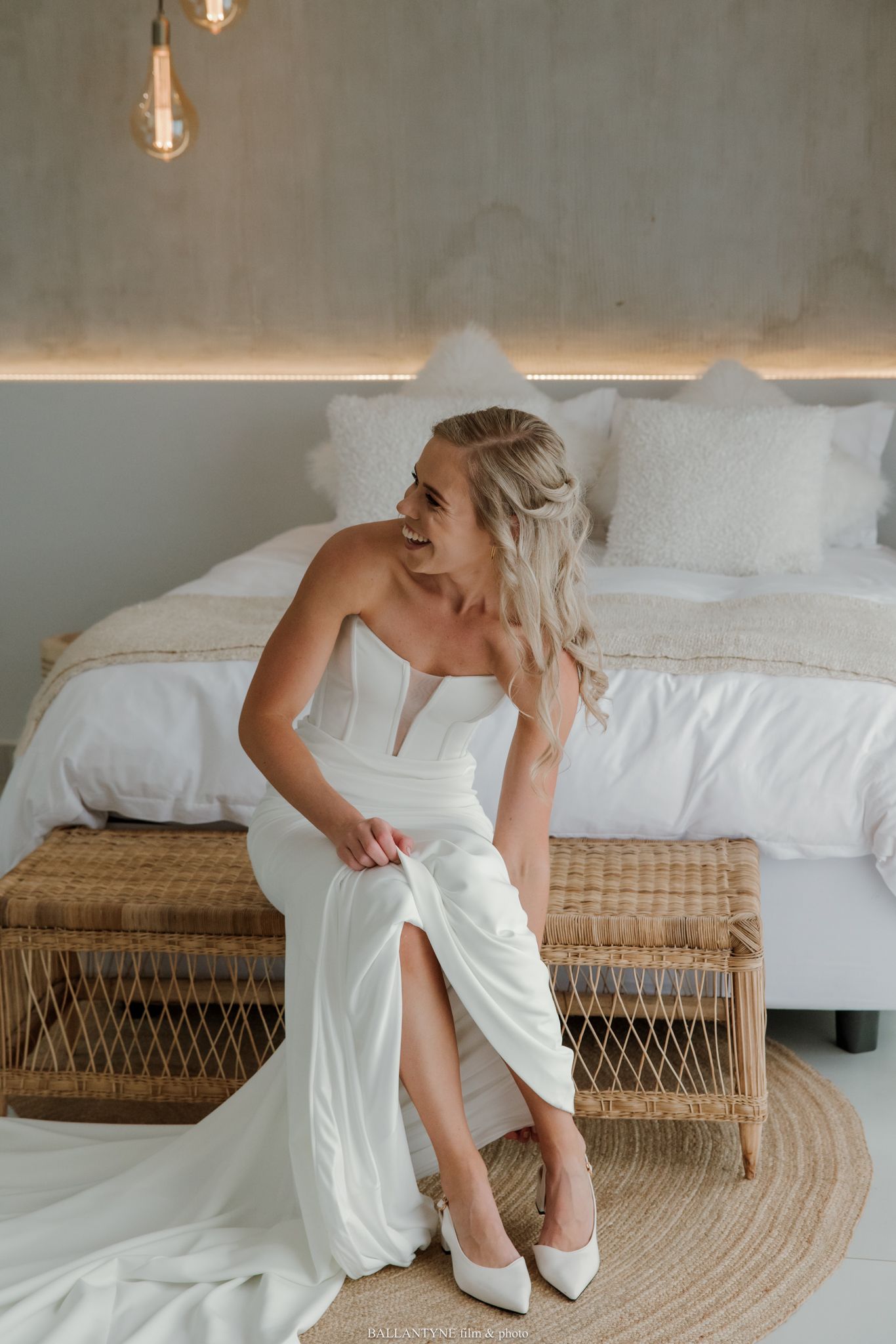 Bride in white strapless wedding gown with slit sits on wicker bench, wearing white heels in elegant bedroom.
