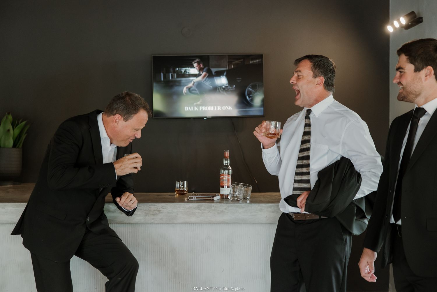 Three men in suits laughing and celebrating at a bar counter with liquor bottles on display.
