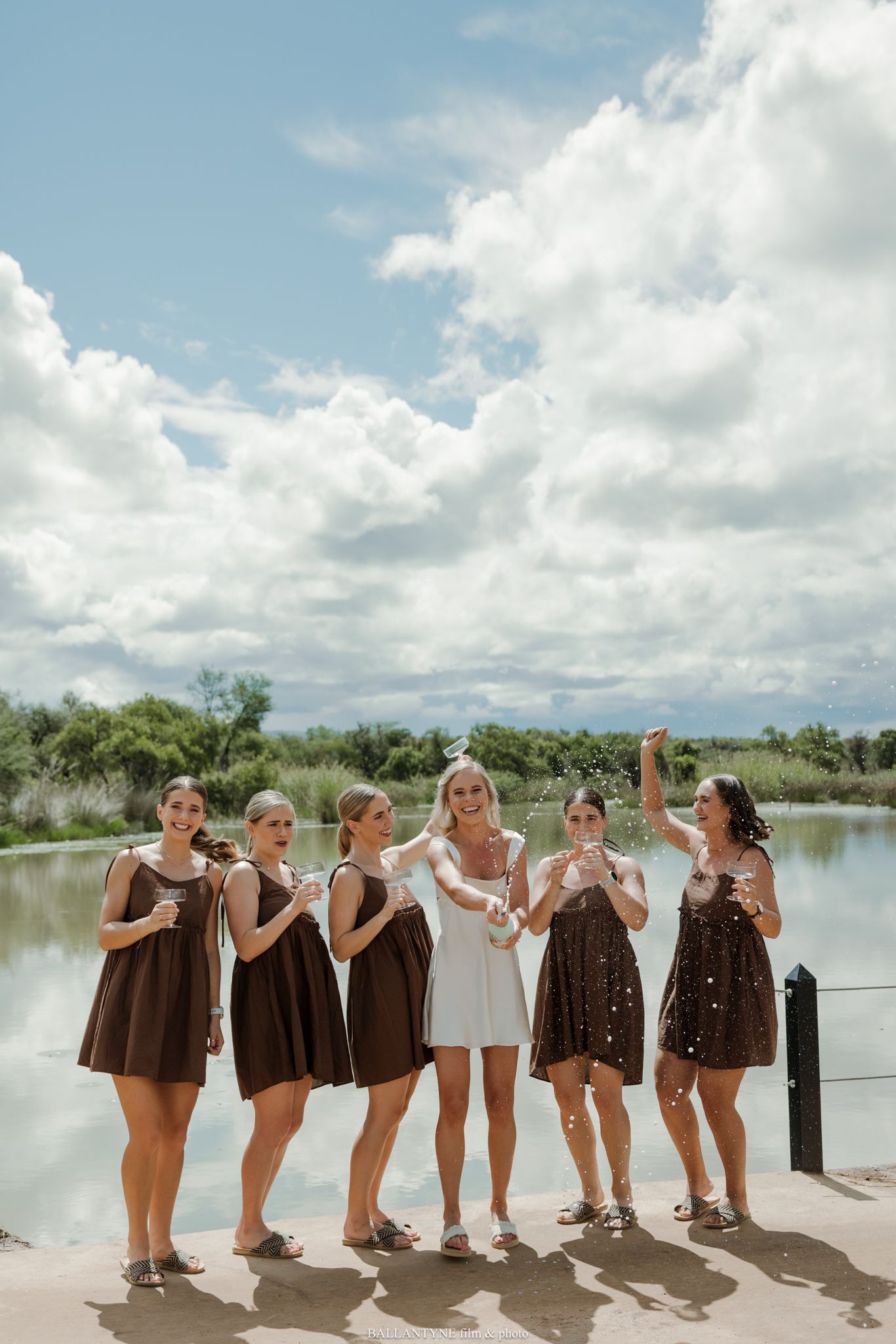Bride in white dress with bridesmaids in brown dresses celebrating with confetti by a lake on a sunny day.
