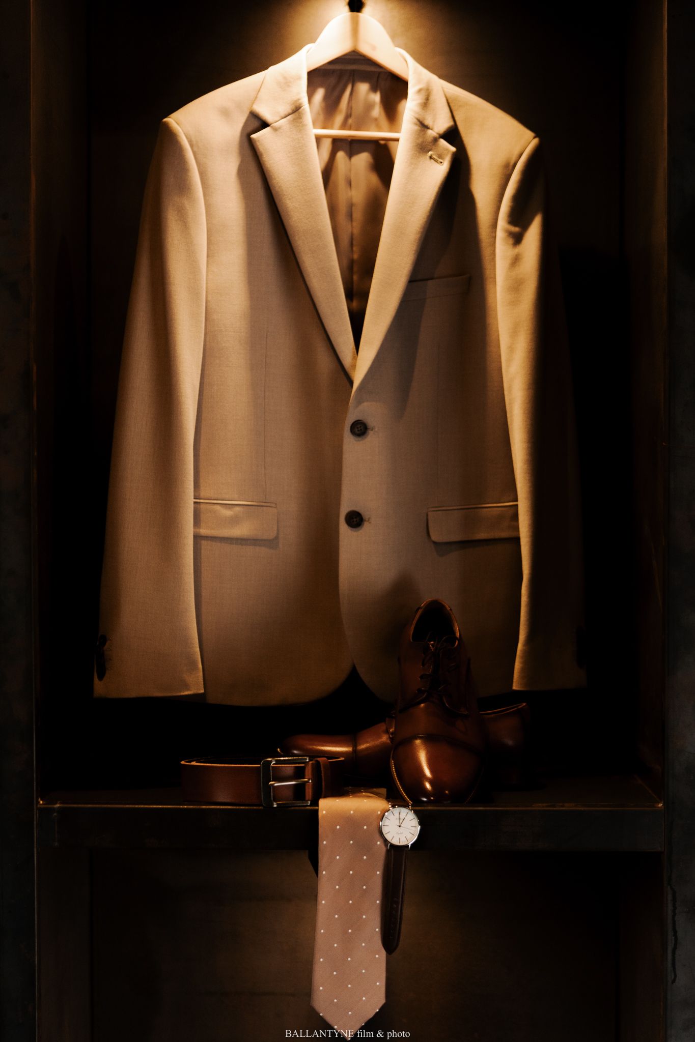 Elegant cream colored suit jacket and tie displayed on a hanger against dark background.