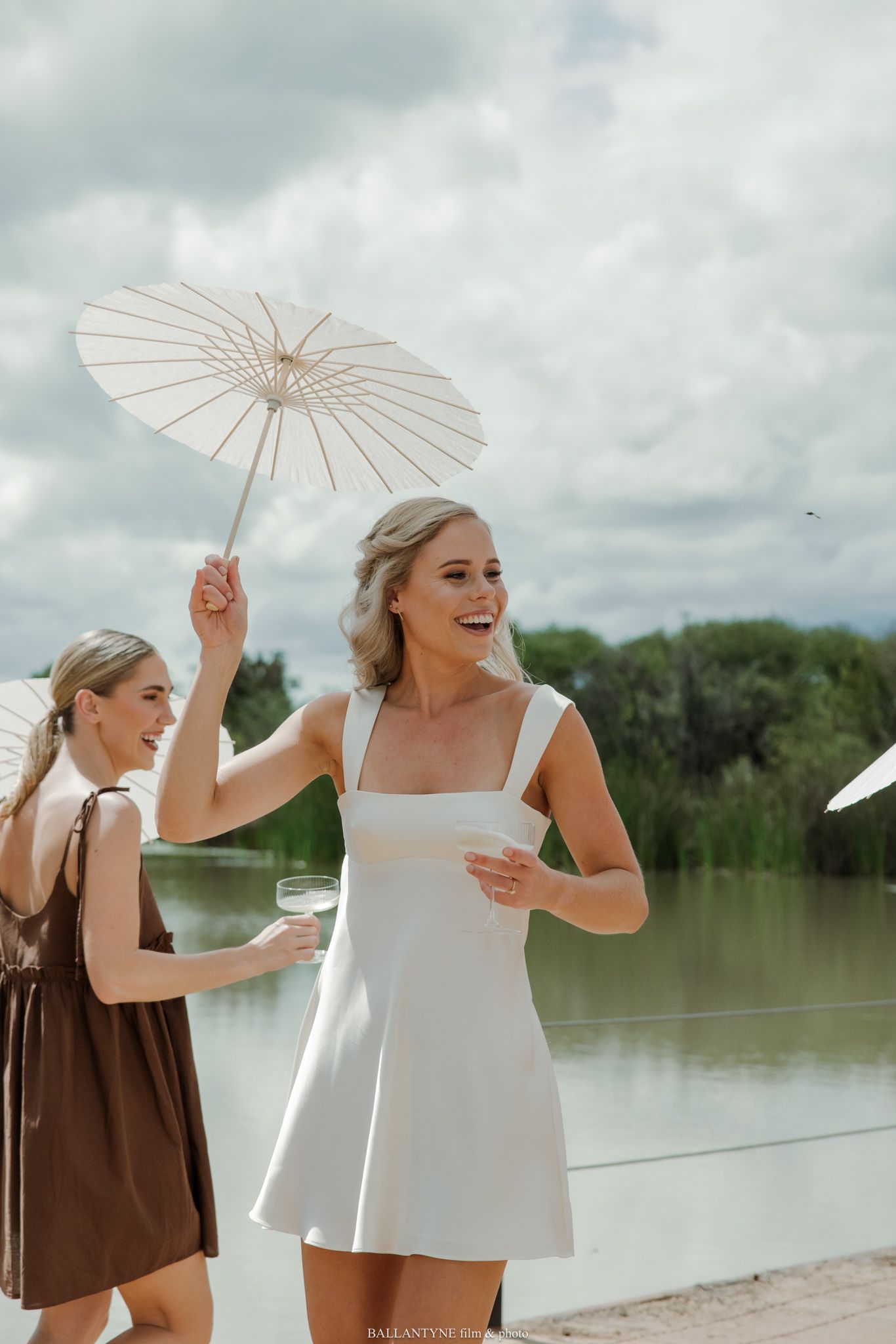 Smiling blonde woman in white mini dress holding parasol umbrella outdoors near lake with friend in brown dress.