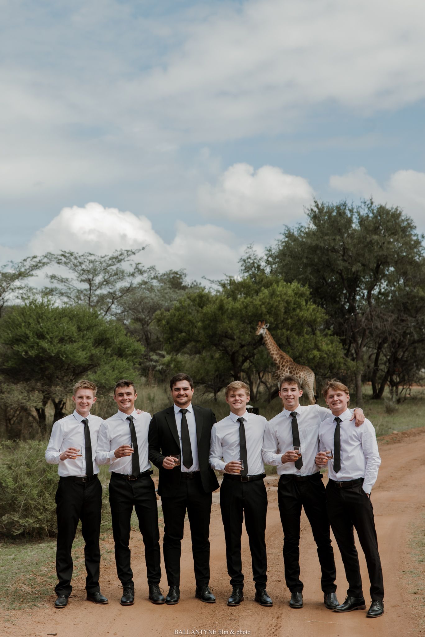 Six groomsmen in white shirts and black ties pose on a dirt road with a giraffe visible in the African bush behind them.