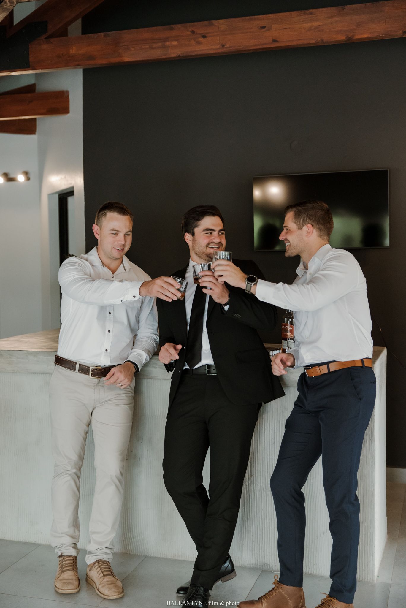 Three men in dress shirts toasting drinks together indoors, with one wearing a black suit, celebrating a special occasion.