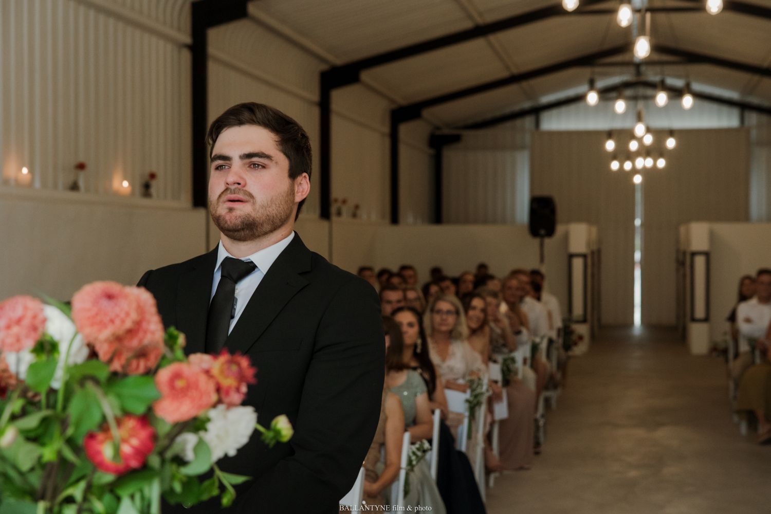 Groom in black suit stands at wedding altar in rustic barn venue with floral arrangements and seated guests.