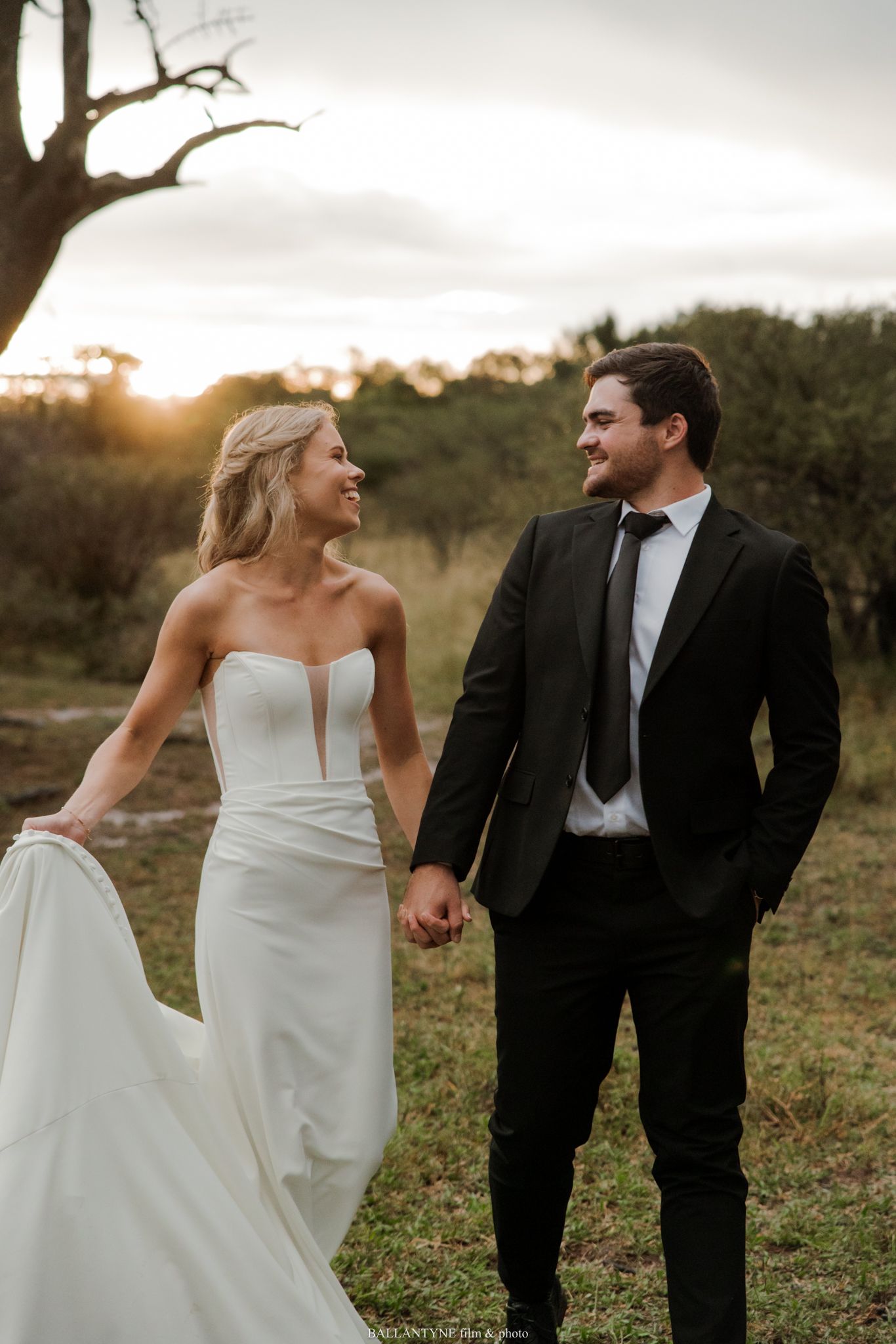 Bride and groom hold hands smiling at each other in a field at golden hour sunset.