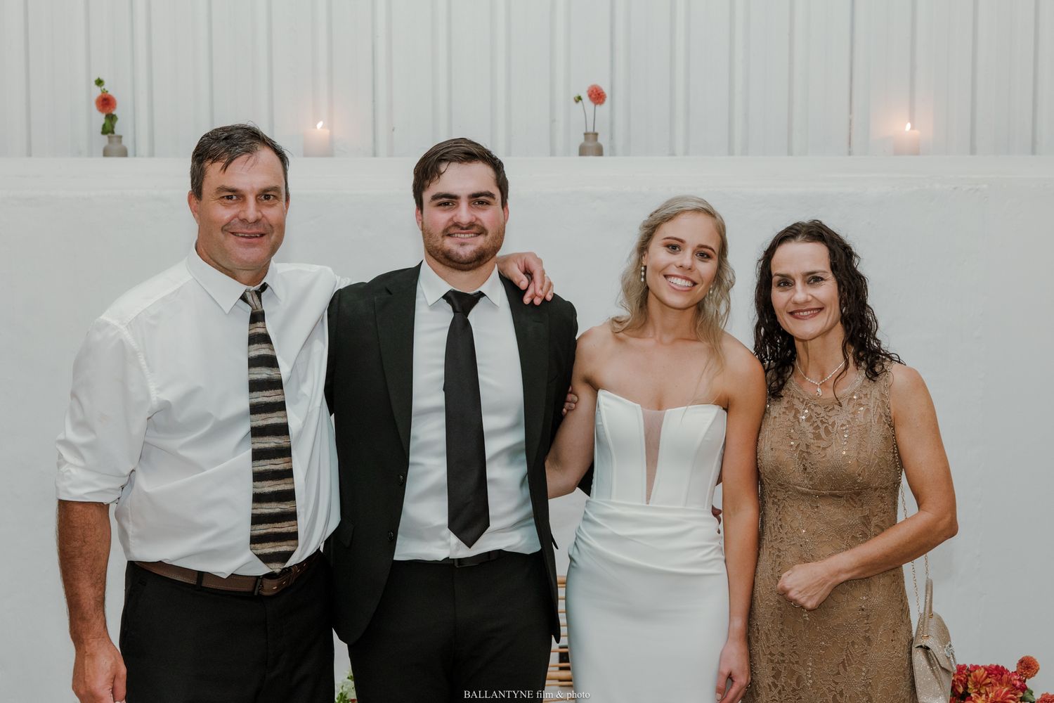 Group of four people posing together at a wedding celebration, with bride in white dress and guests in formal attire.