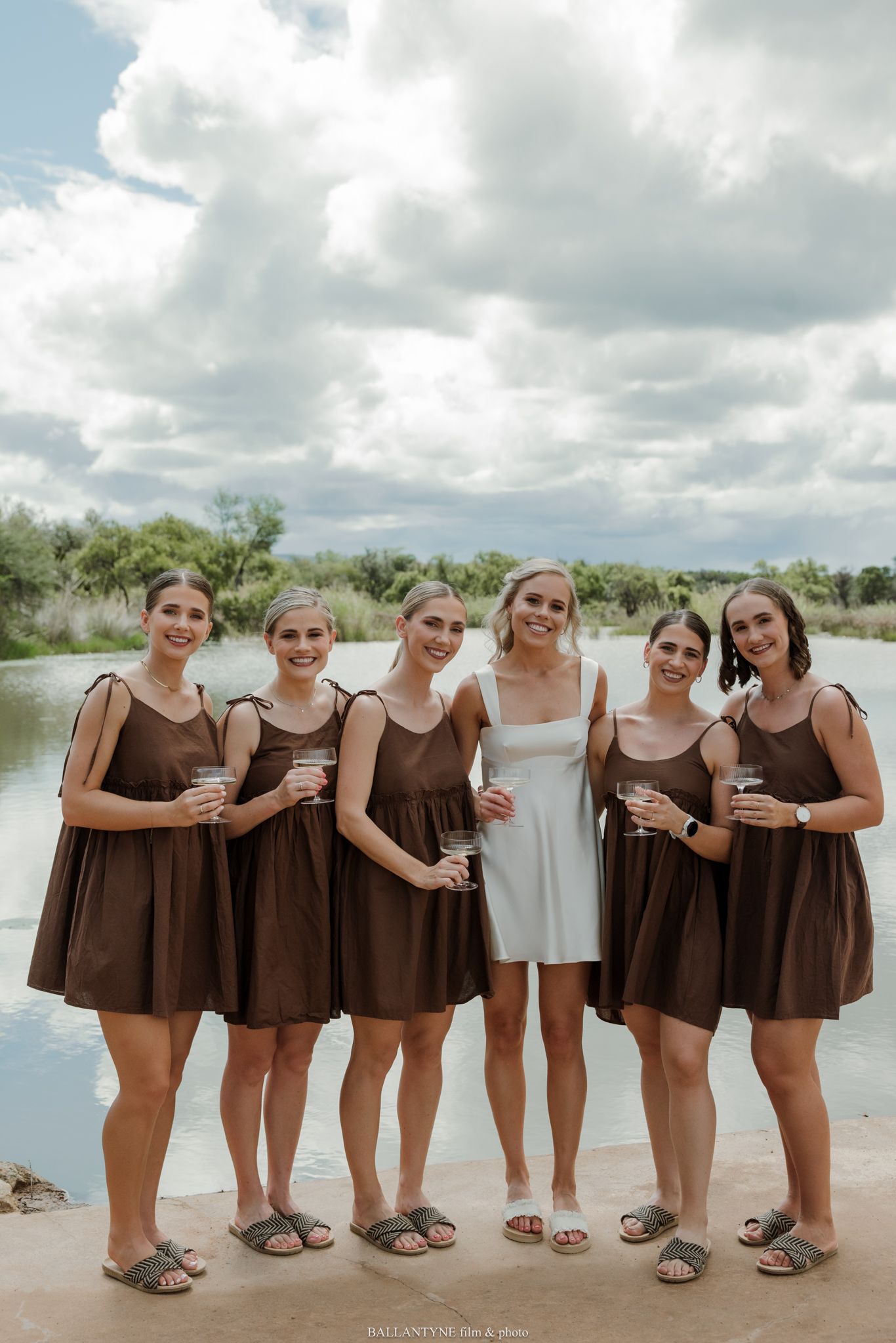 Bride in white dress with five bridesmaids in brown mini dresses posing outdoors near a river under cloudy skies.