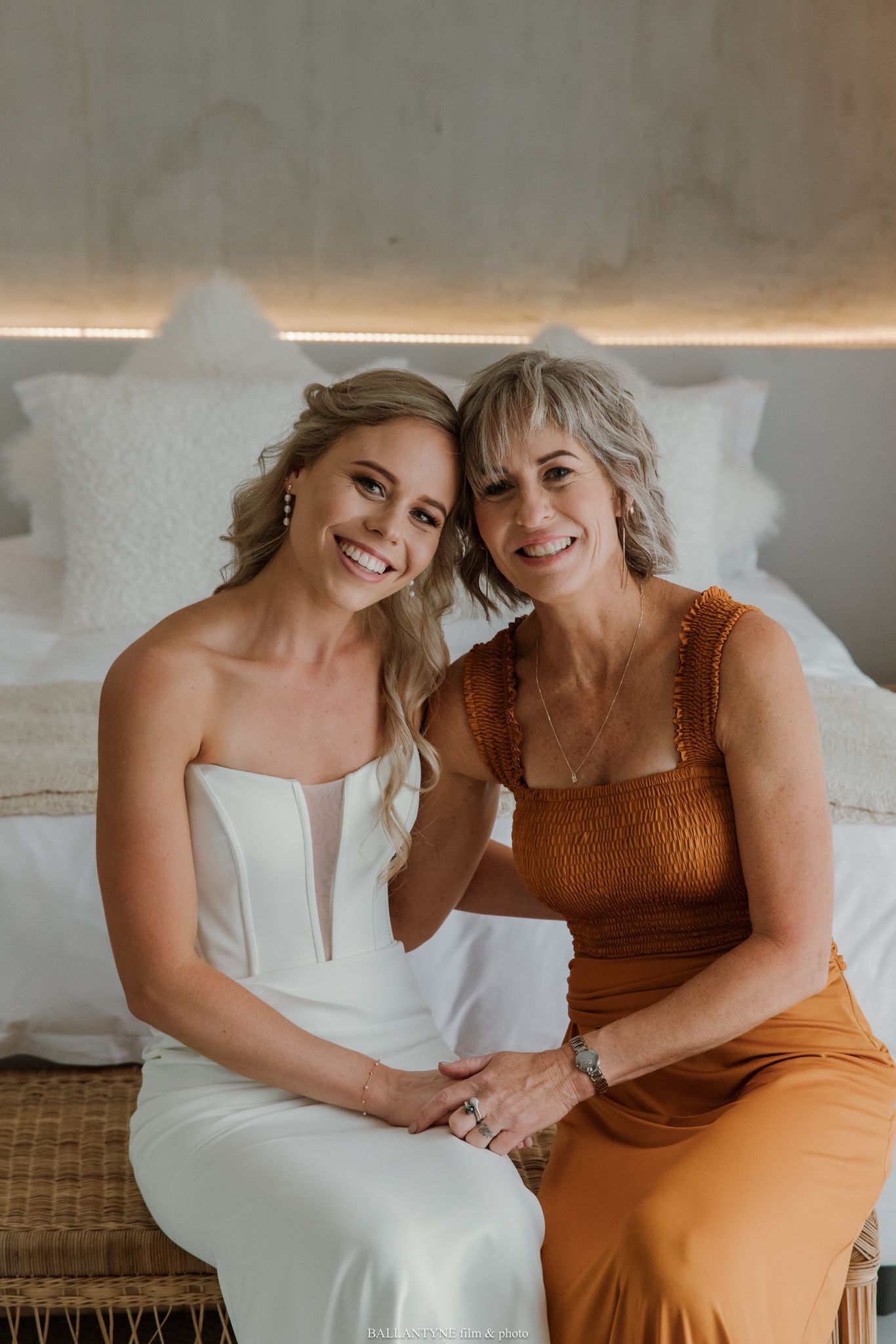 A bride in a white gown sits with an older woman in an orange dress, smiling together on a bed.