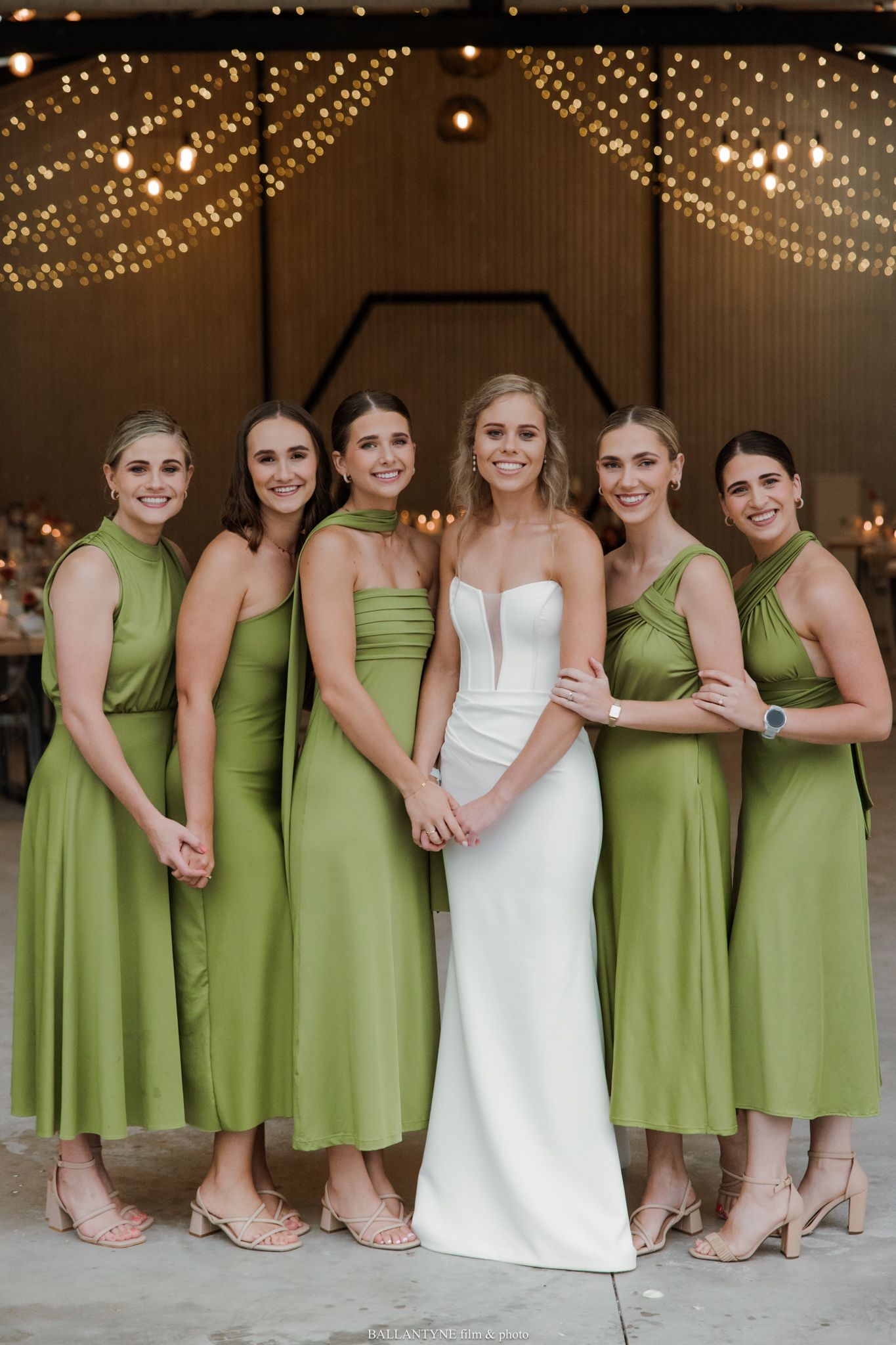 Bride in white gown with five bridesmaids in olive green dresses posing in a barn venue with string lights.