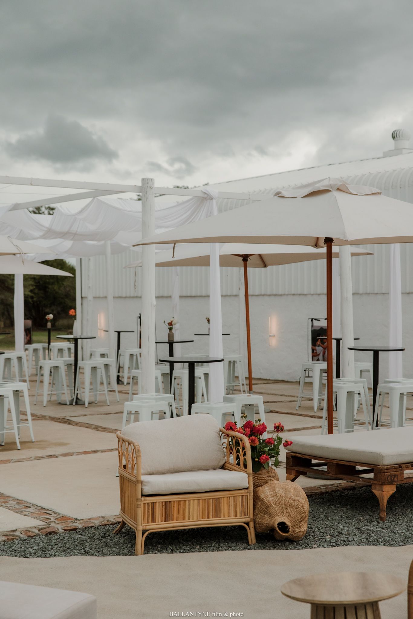 Elegant outdoor event space with white draping, rattan furniture, and patio umbrellas under dramatic cloudy skies.