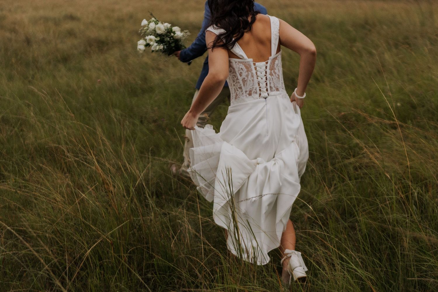 Bride in white lace wedding dress running through green meadow holding bouquet of white flowers.