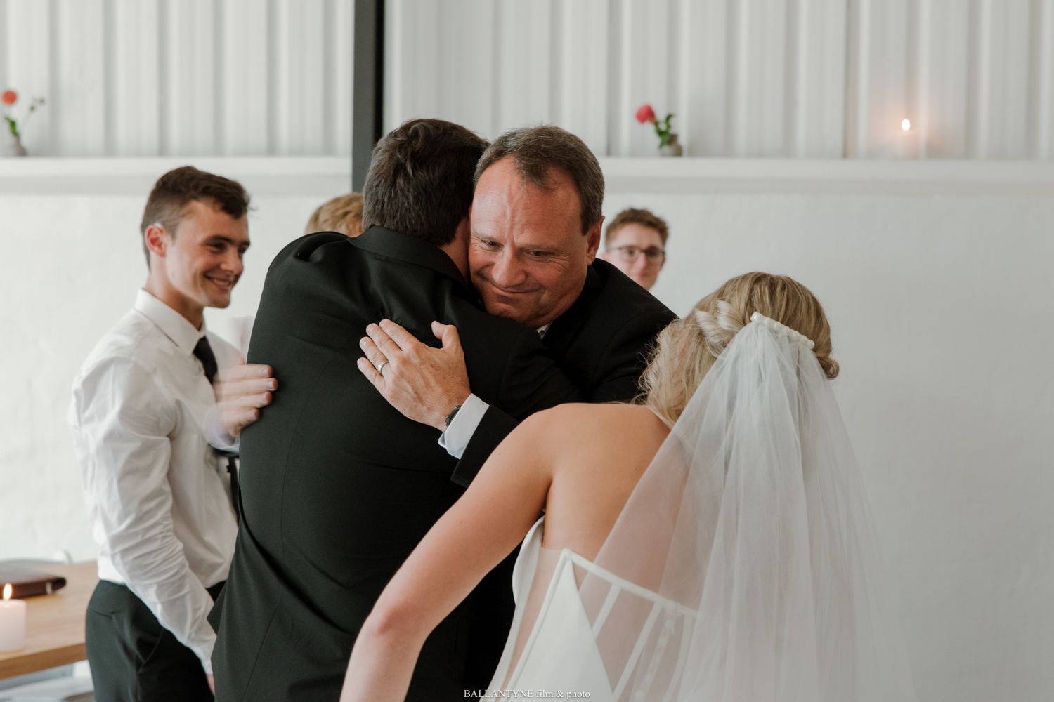 Emotional wedding hug between groom and guest, bride in white veil nearby, guests watching in bright venue.