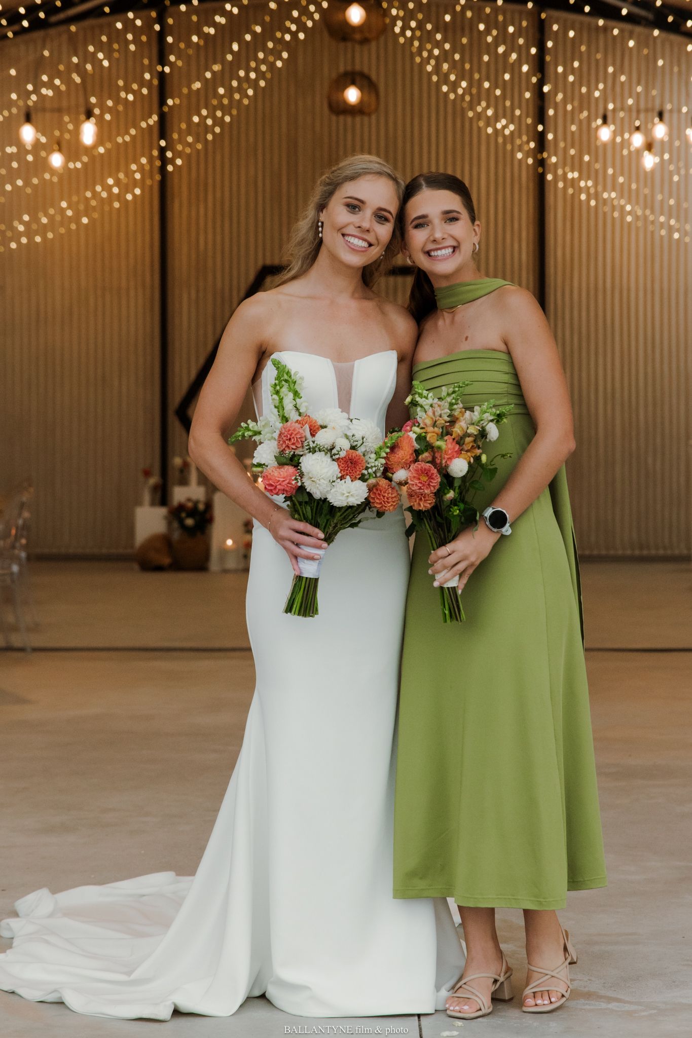 Bride in white gown and bridesmaid in green dress holding floral bouquets, smiling together at a wedding venue.