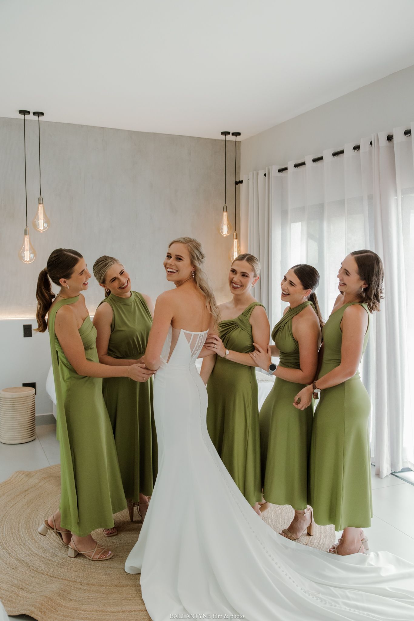 Bride in white gown with five bridesmaids in olive green dresses laughing together in a bright modern room.