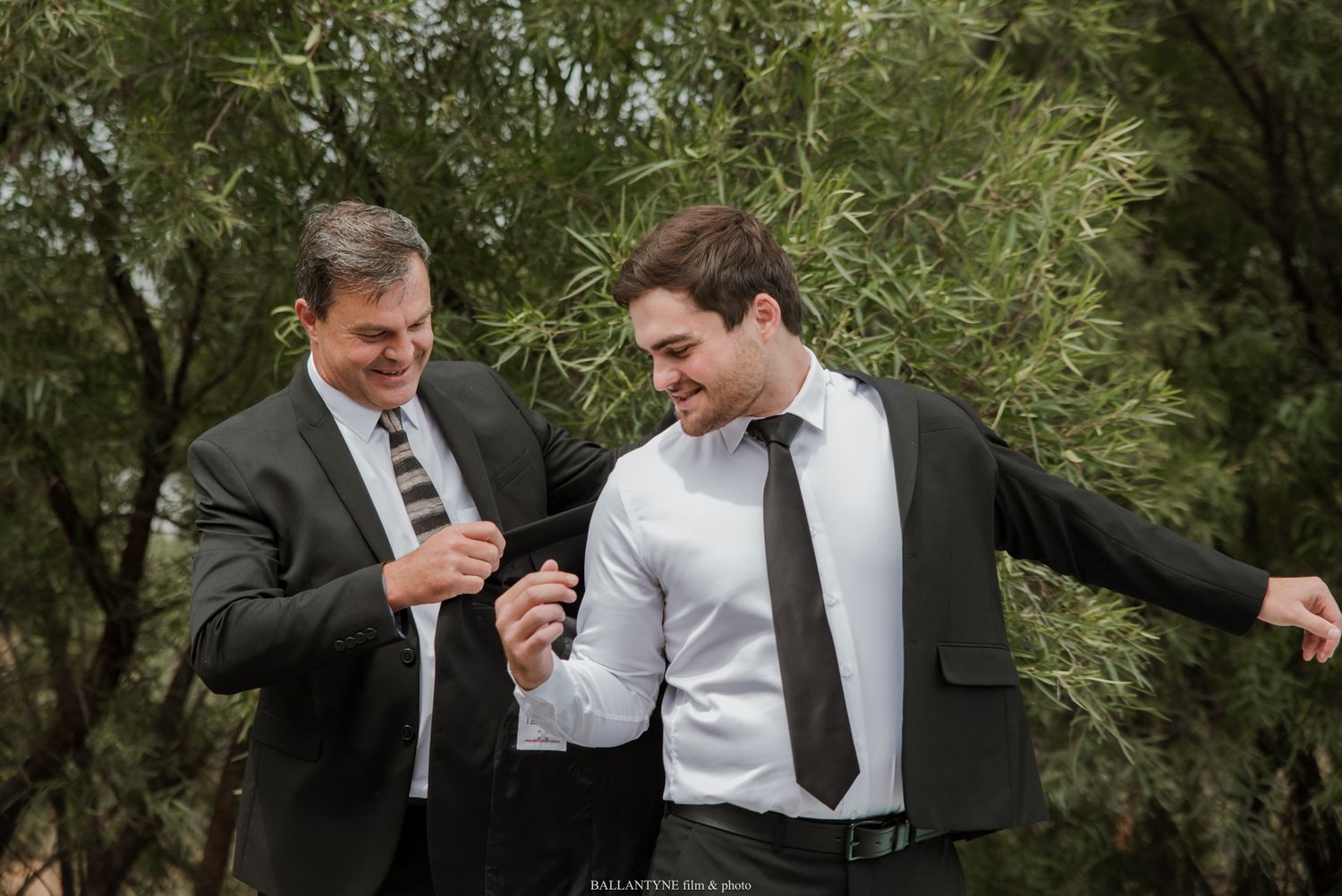 Two men in dark suits laugh together outdoors surrounded by lush green trees at a formal event.