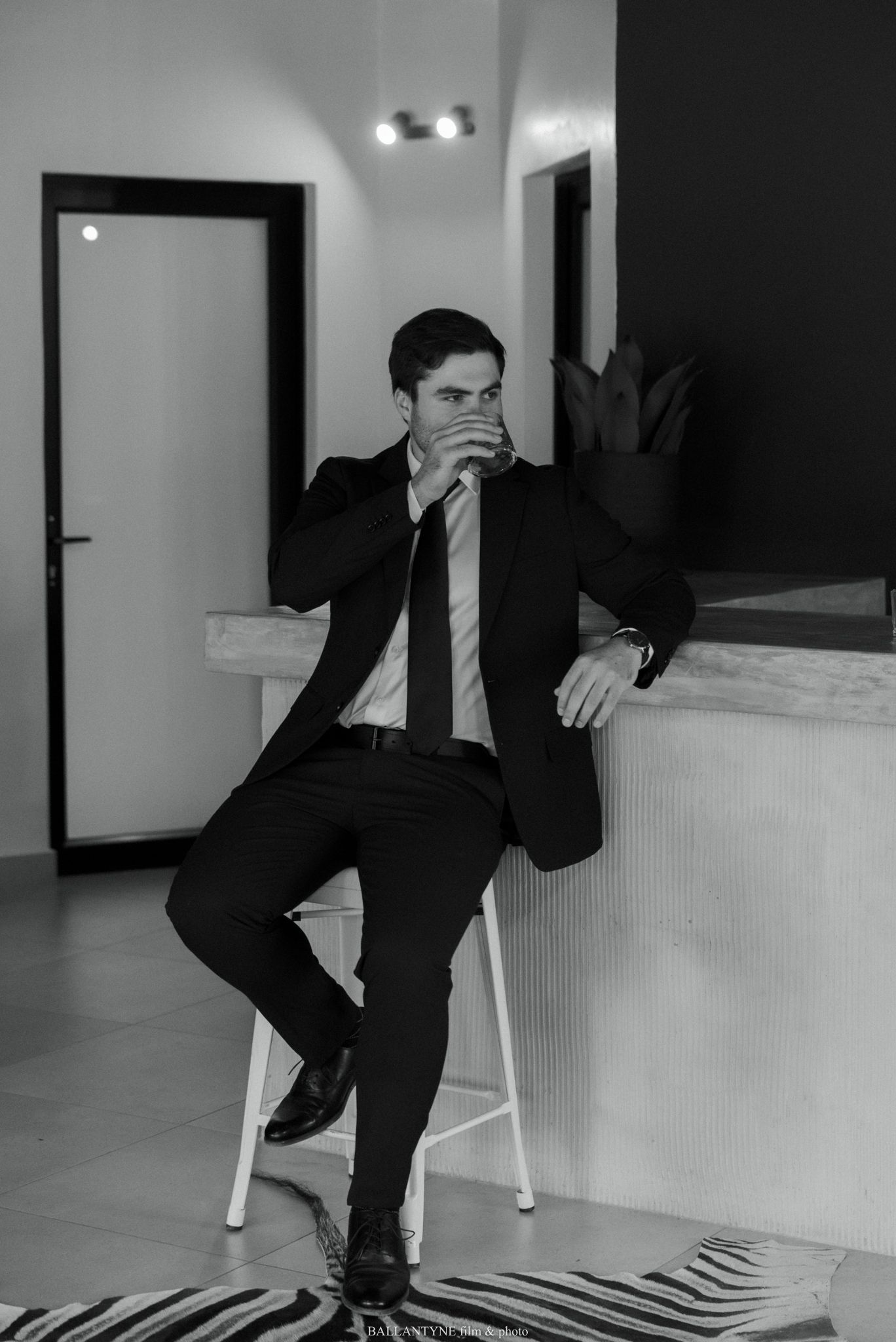 A well-dressed man in a black suit and tie sits on a stool in a modern interior, resting his chin on his hand.