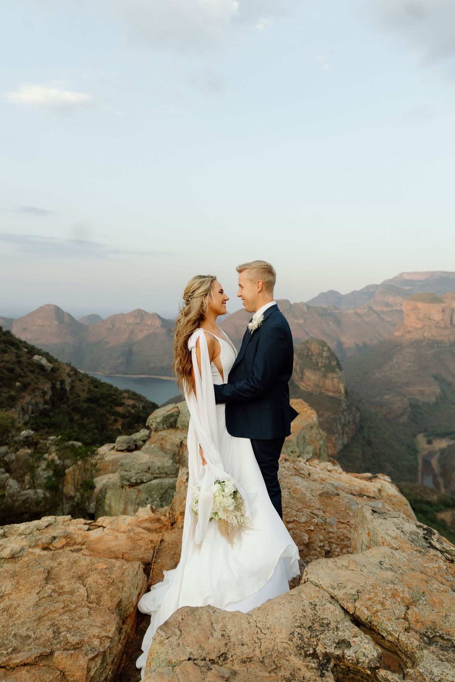 Wedding couple stands on mountain cliff edge with dramatic landscape views.