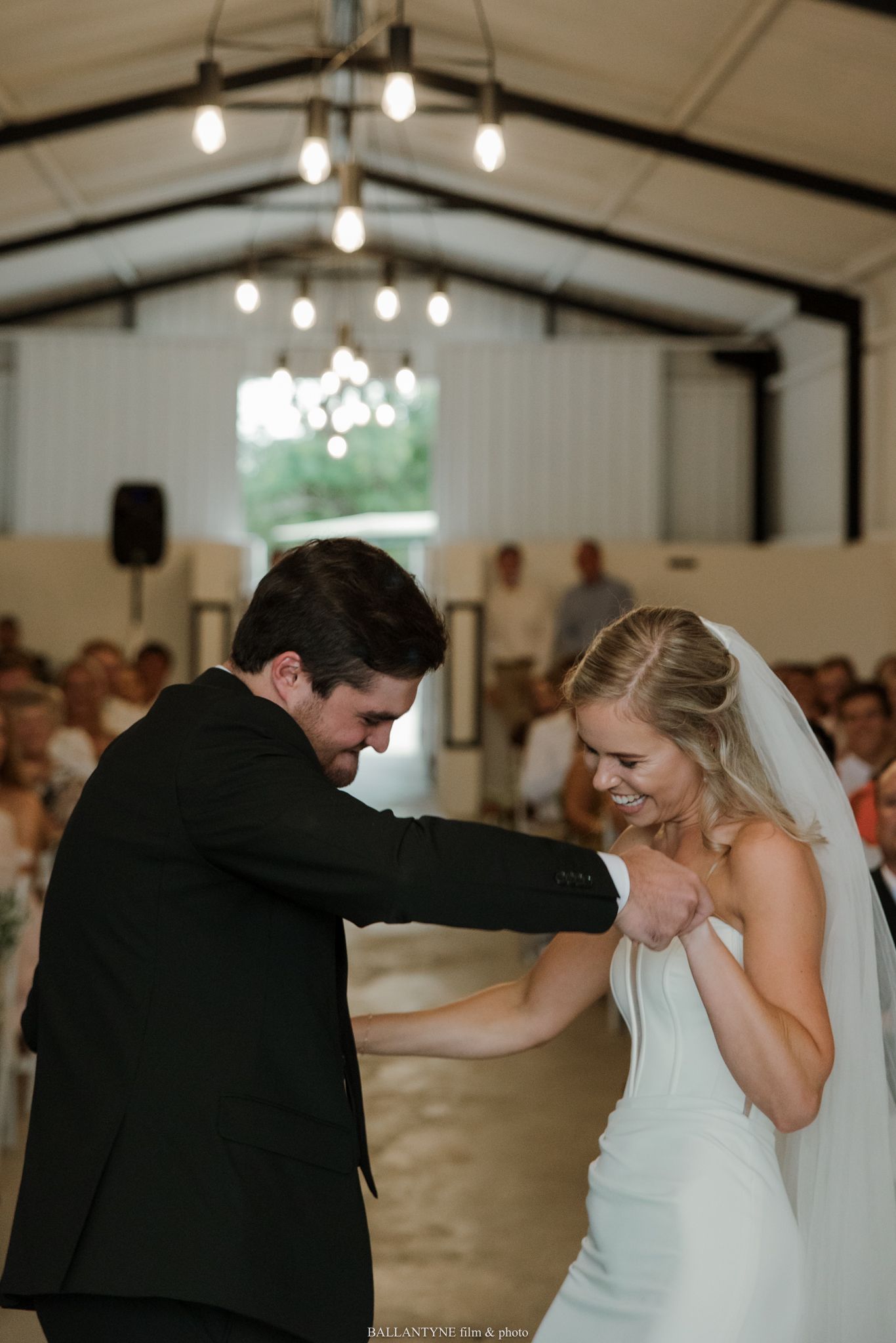 Bride and groom share their first dance in a white barn wedding venue with warm Edison bulb lighting.