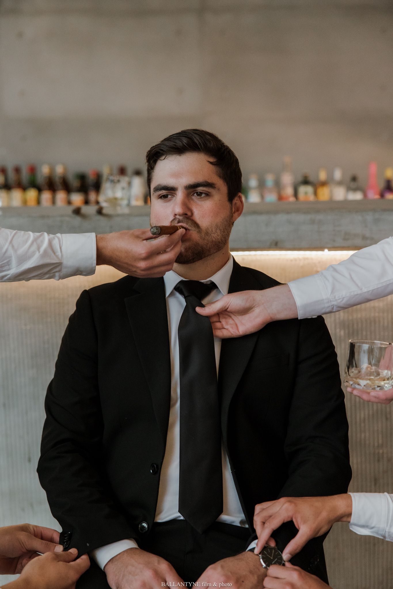 Groom in black suit being groomed by friends at a bar, having his tie adjusted and cigar held.
