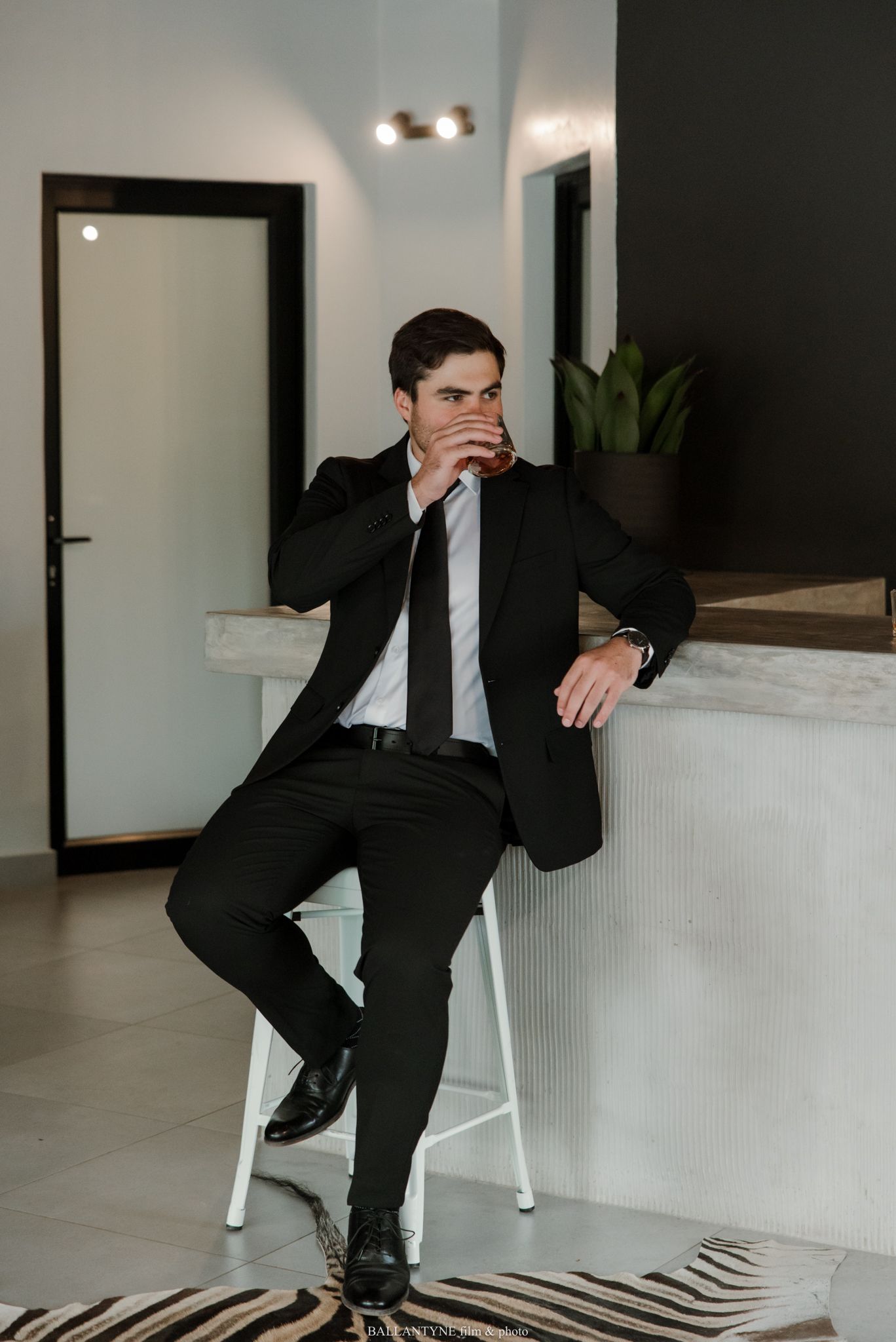 A well-dressed man in a black suit and tie sits on a white stool, resting his arm on a counter in a modern interior.