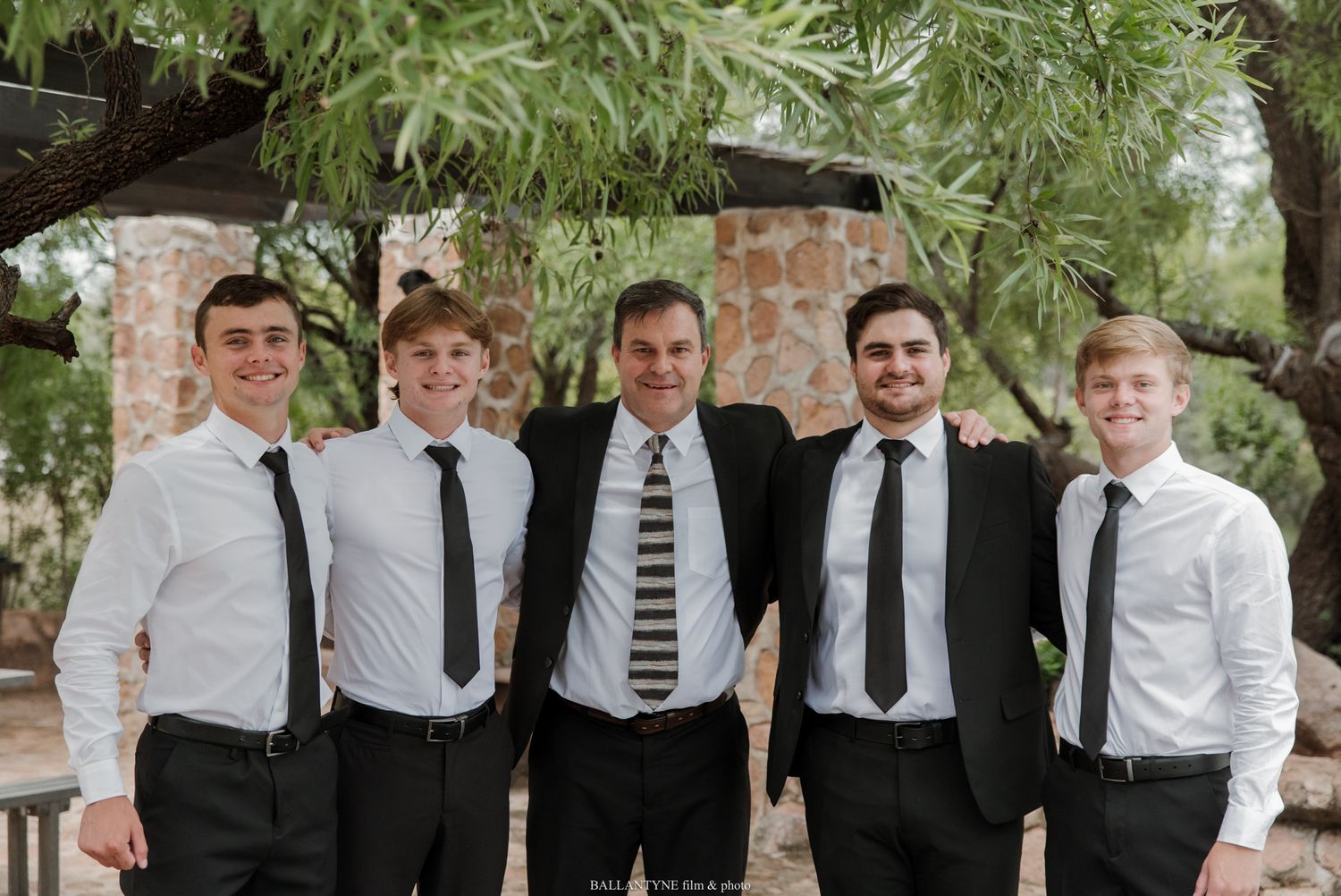 Five men in white shirts and black ties smile together outdoors under trees at a formal event.