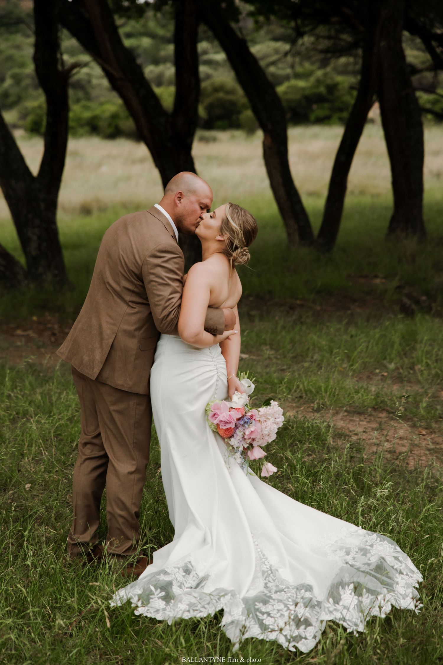 A romantic wedding photo shows a couple kissing in a wooded area, with the bride holding pink flowers and wearing a lace dress.