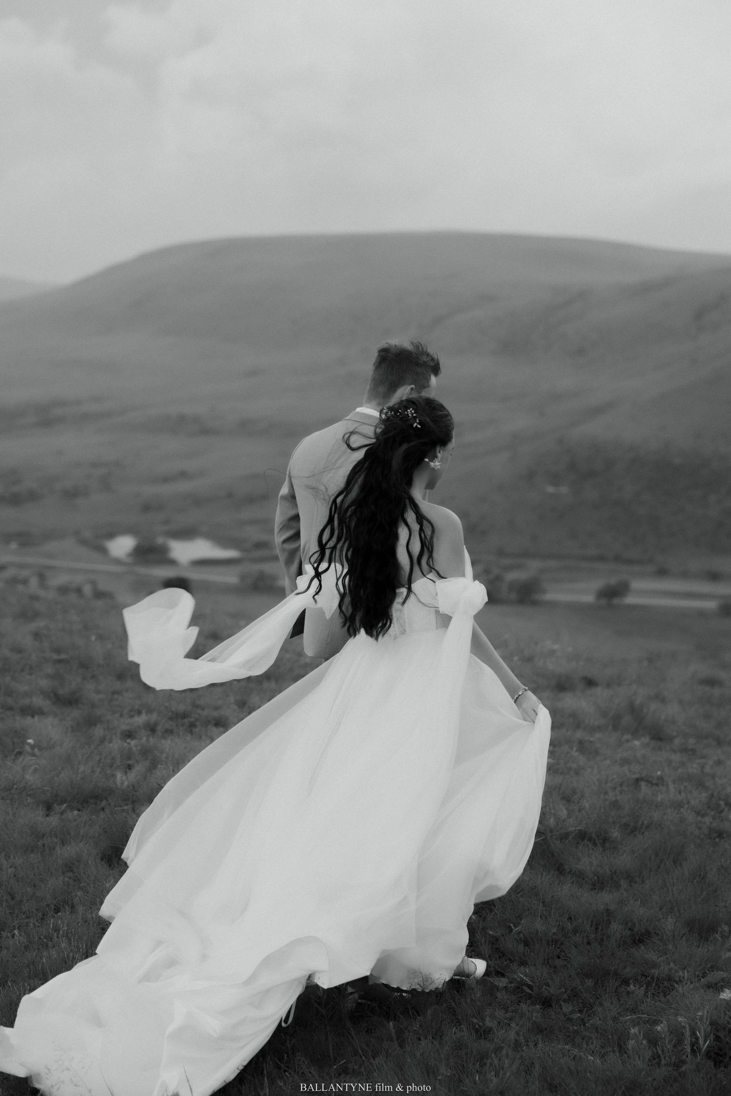 Black and white photo of a couple embracing on a hilltop with flowing wedding dress in the wind.