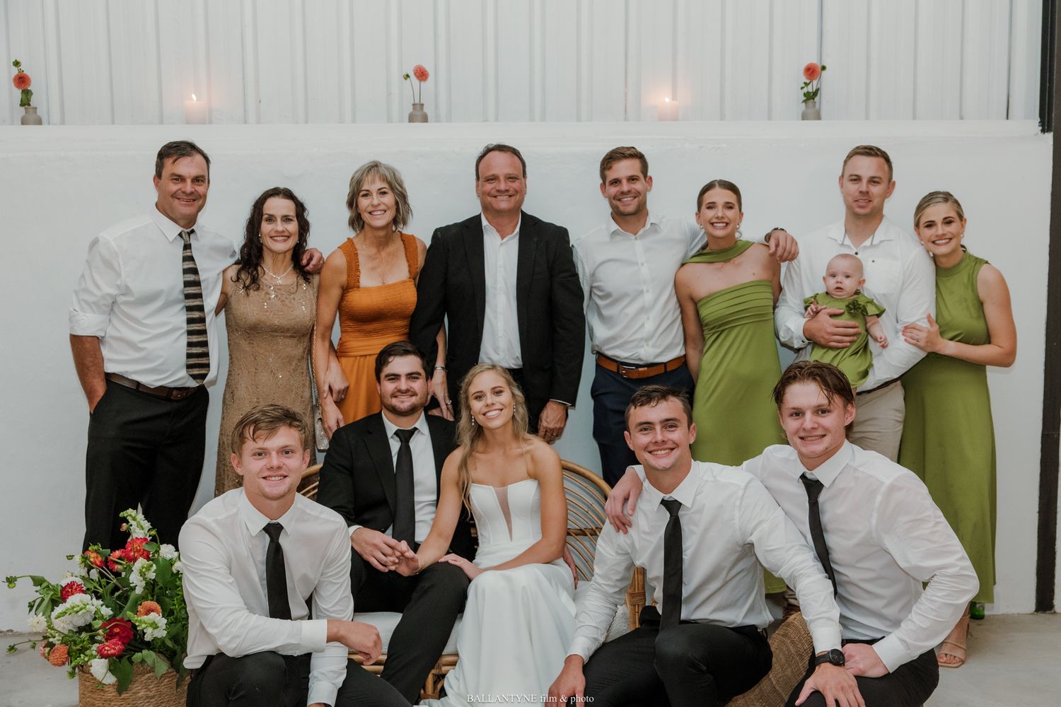 Wedding group photo with bride in white gown surrounded by family and friends in formal attire at elegant venue.