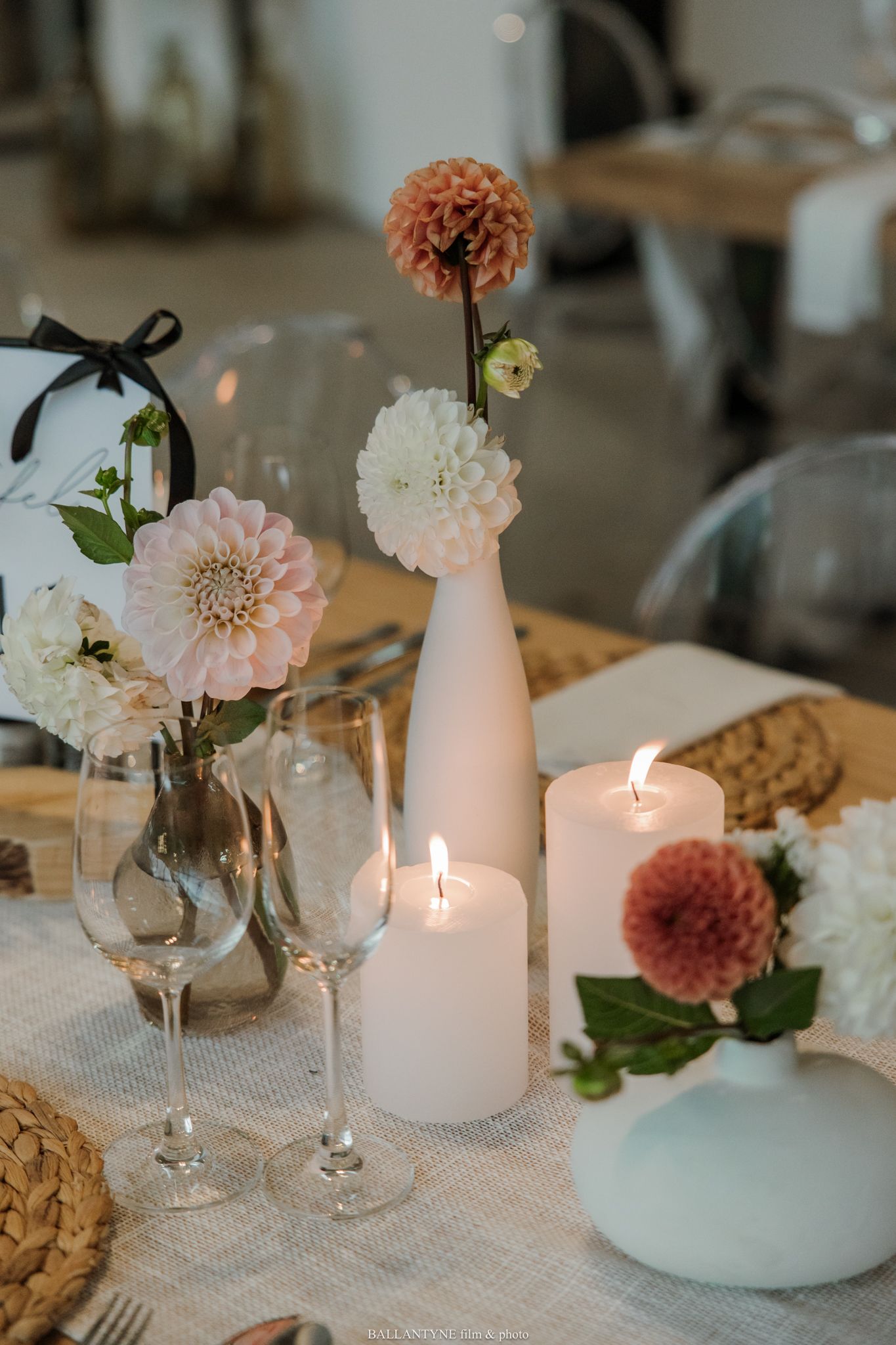 Wedding table decor with pink and coral dahlias in white vases, lit candles, and crystal wine glasses.