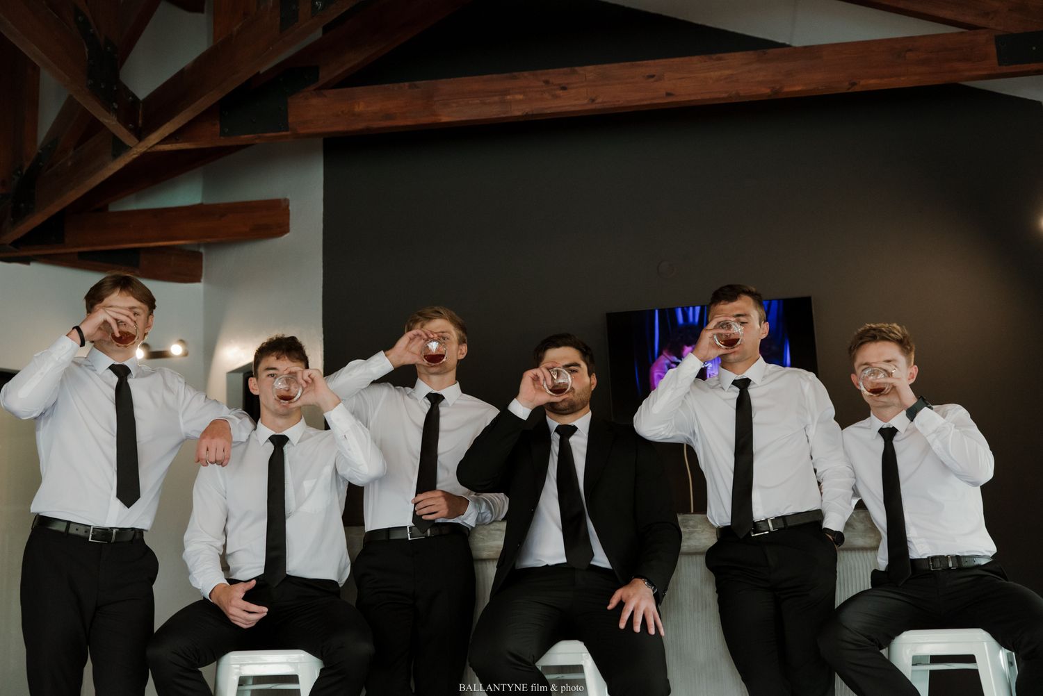 Six groomsmen in white shirts and black ties strike funny poses making mustache and glasses gestures at a wedding venue.