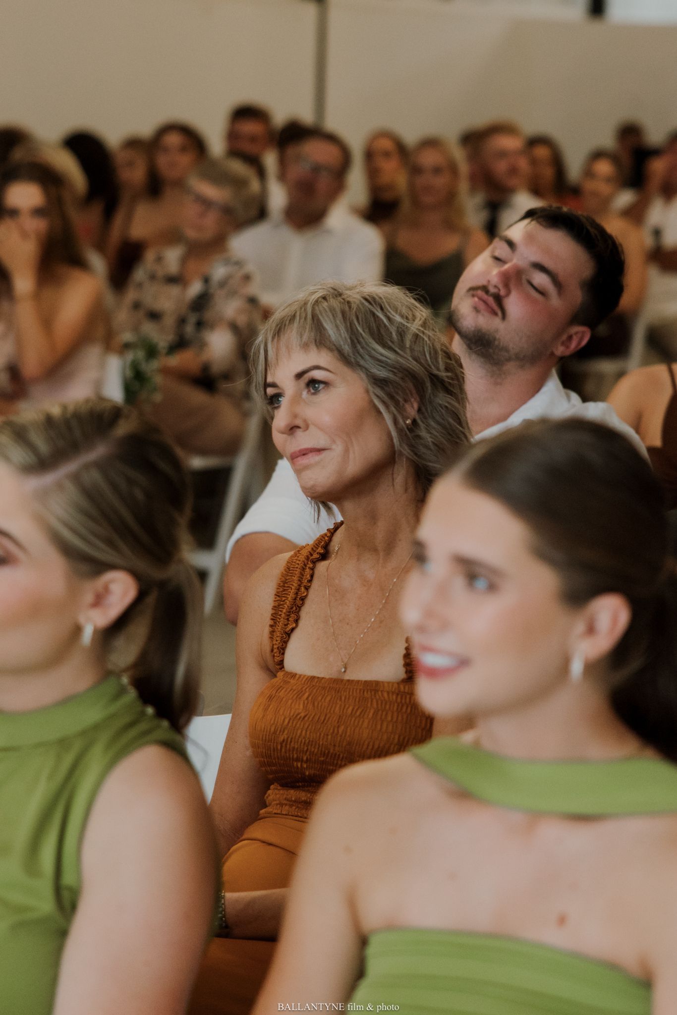 Wedding guests seated in rows, women in green dresses and a woman in brown dress watching a ceremony.