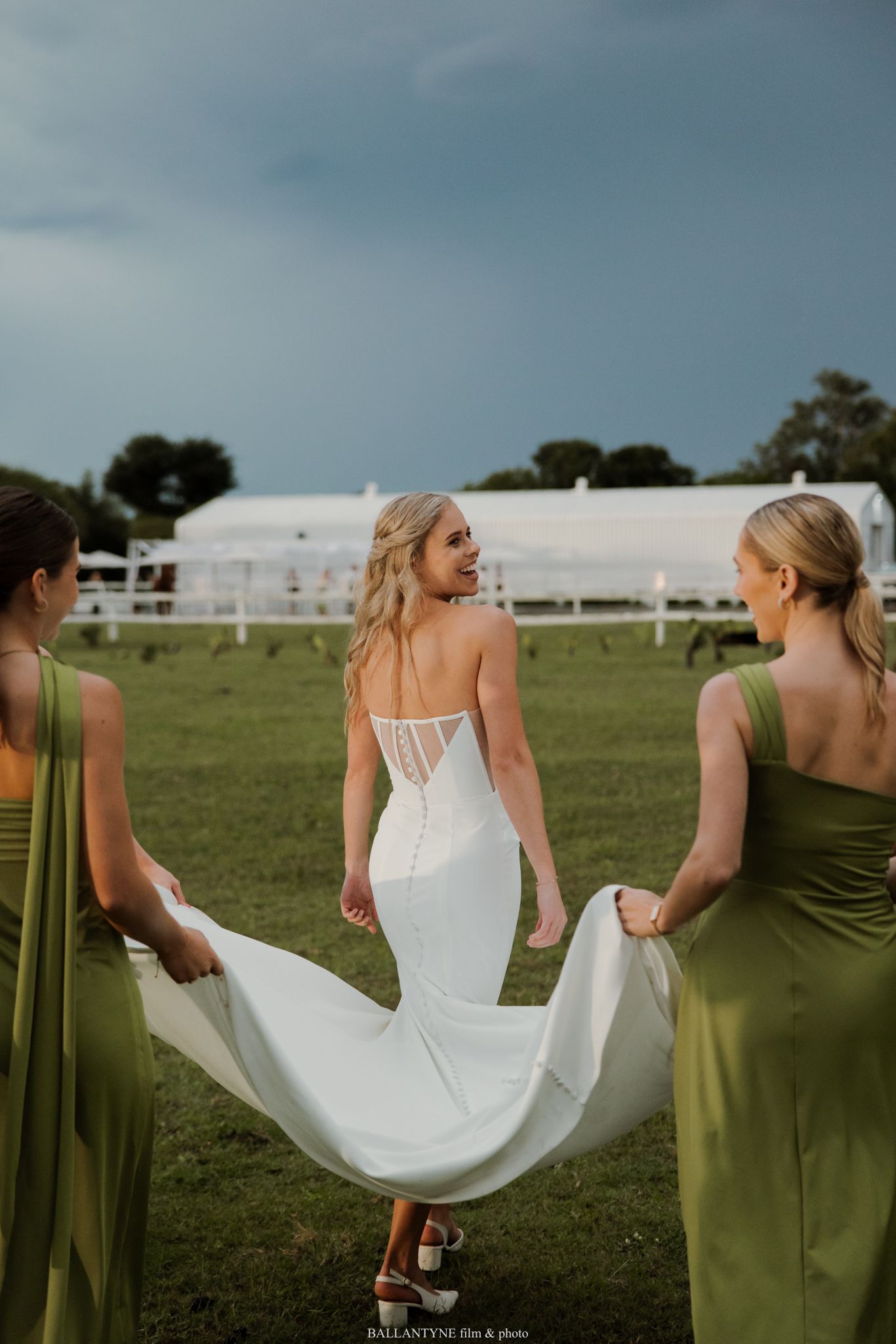 Bride in white lace gown walks across green field as bridesmaids in olive dresses carry her train.