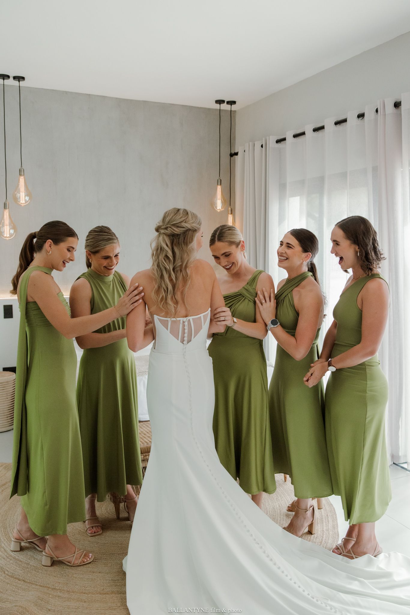 Bride in white lace gown surrounded by five bridesmaids in olive green dresses during wedding preparations.
