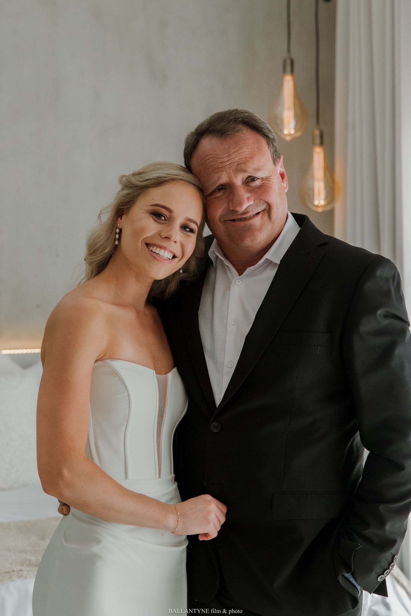 Bride in strapless white gown smiles with older man in black suit at elegant wedding venue.