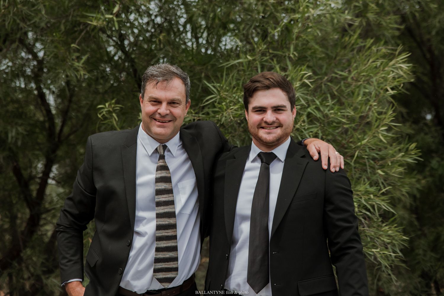 Two men in suits smiling together outdoors at a wedding celebration.