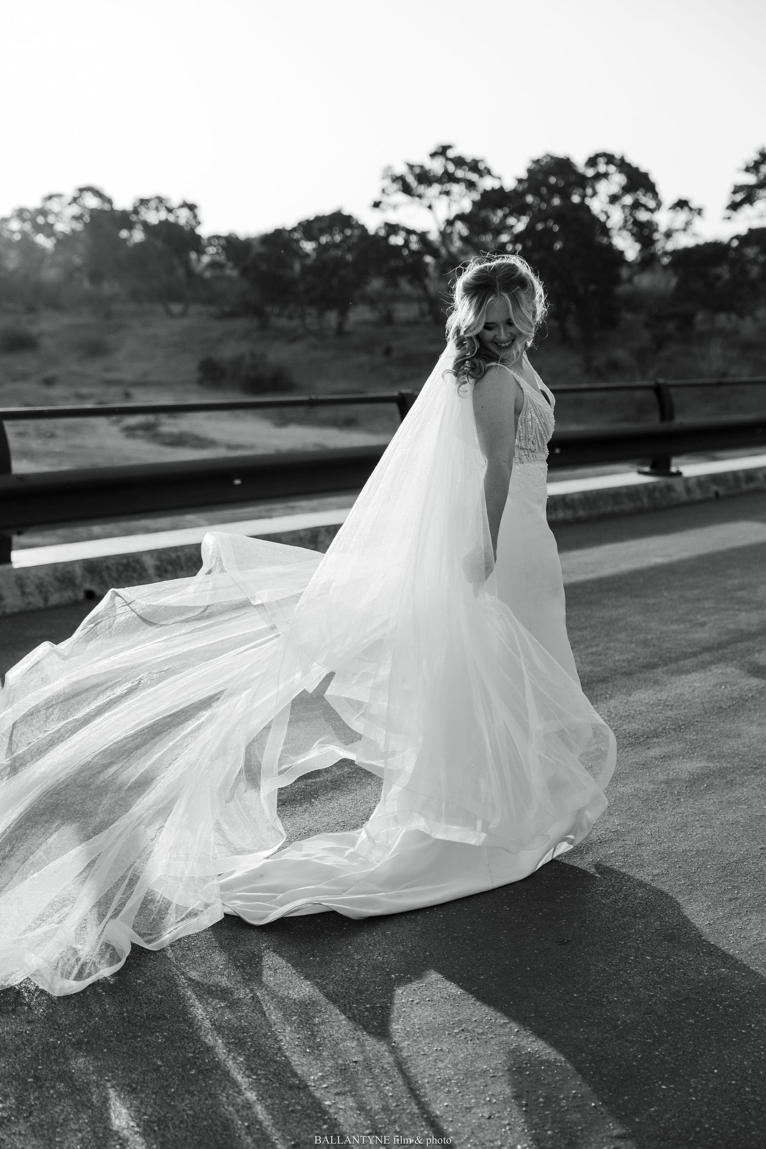 A flowing white wedding dress and veil catch the wind dramatically in black and white against a rural landscape.
