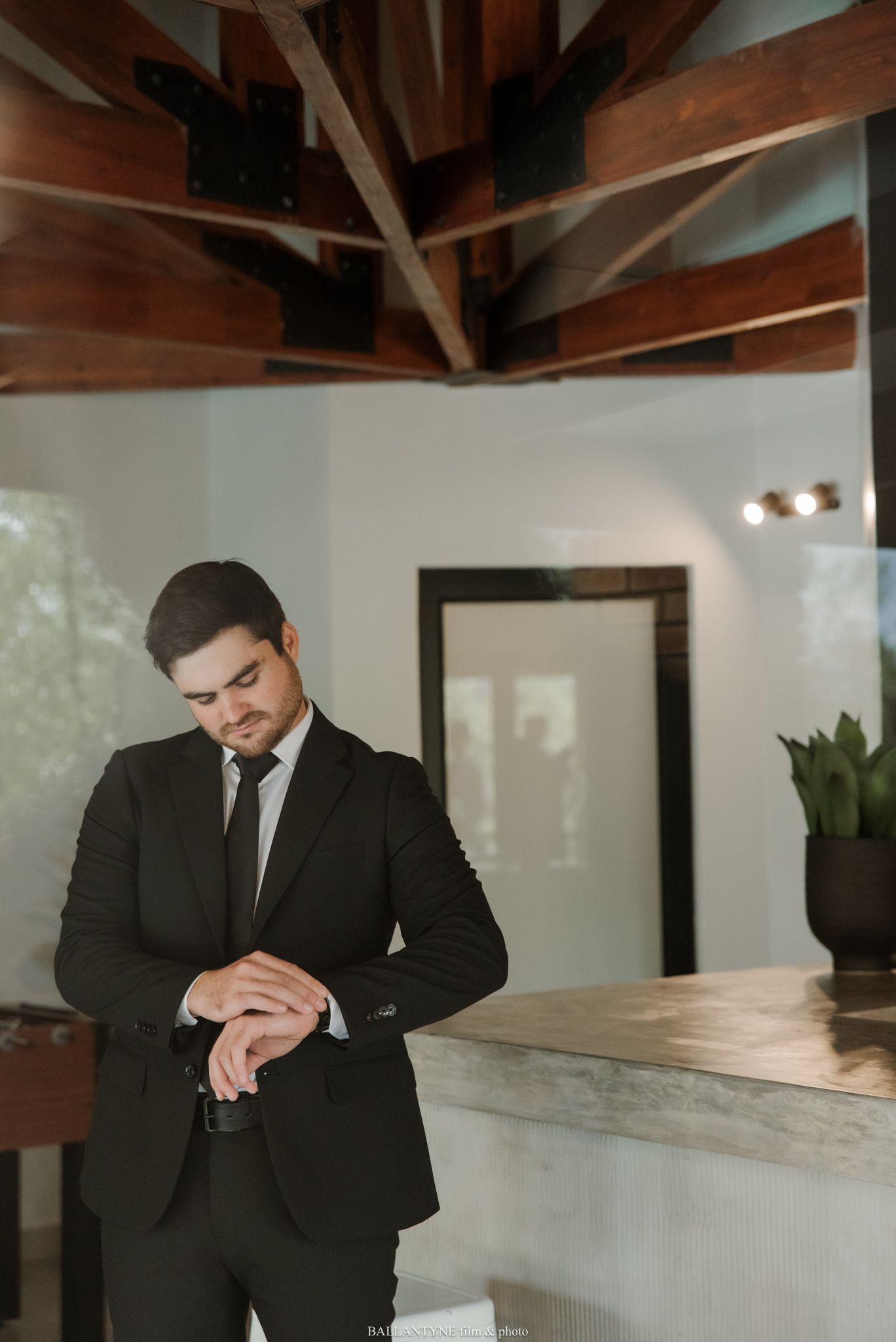 Groom in black suit adjusting cufflinks before wedding ceremony in elegant venue with wooden beam ceiling.
