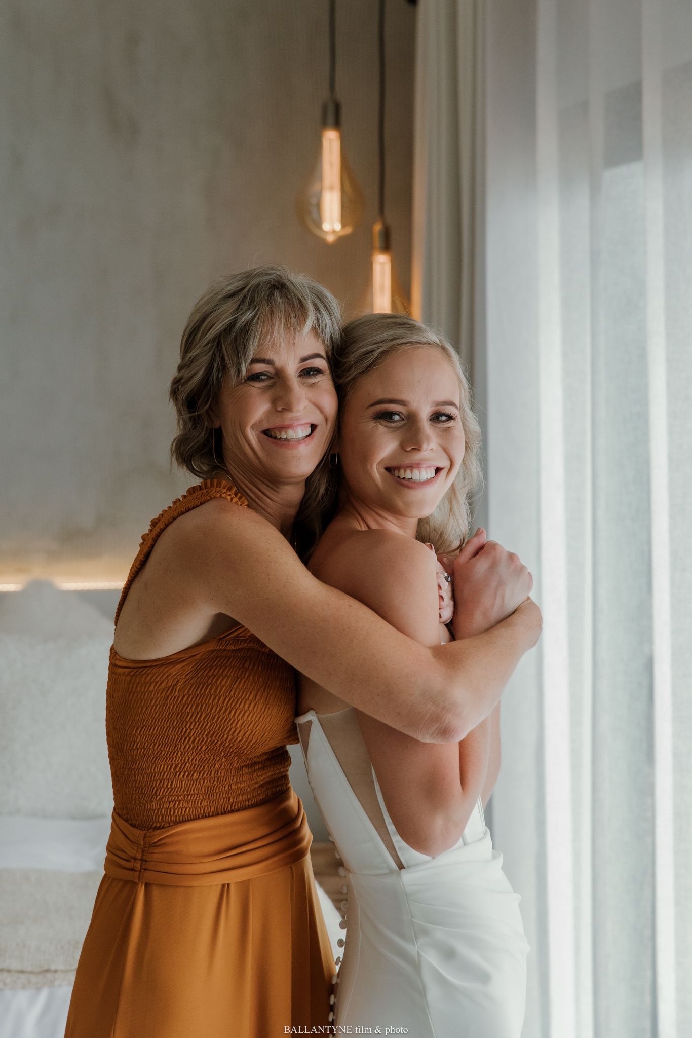 A bride in a white gown embraces an older woman in an orange dress, both smiling warmly near a bright window.