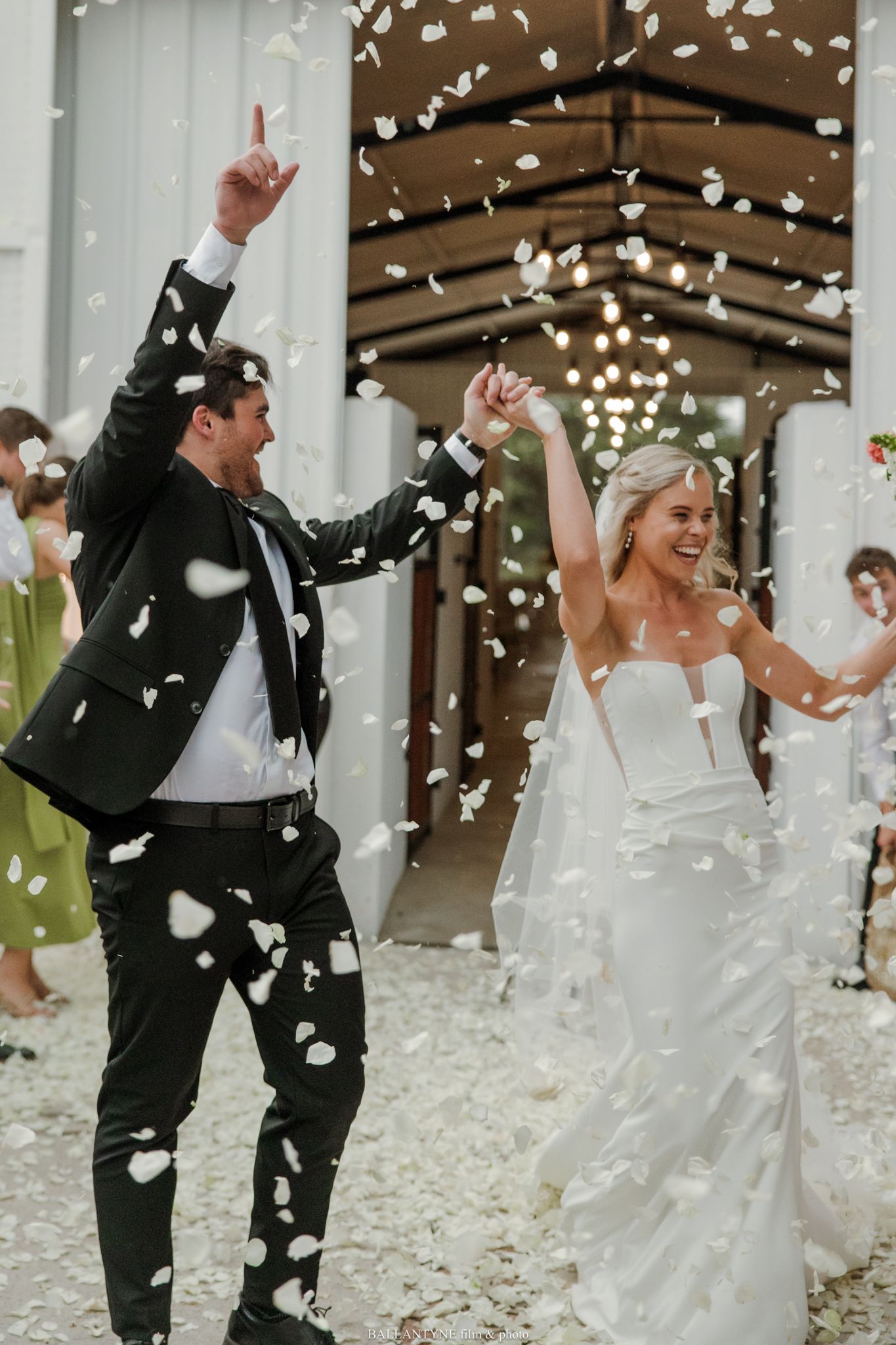 Joyful newlyweds celebrate their wedding exit with white flower petals raining down as guests cheer in the background.