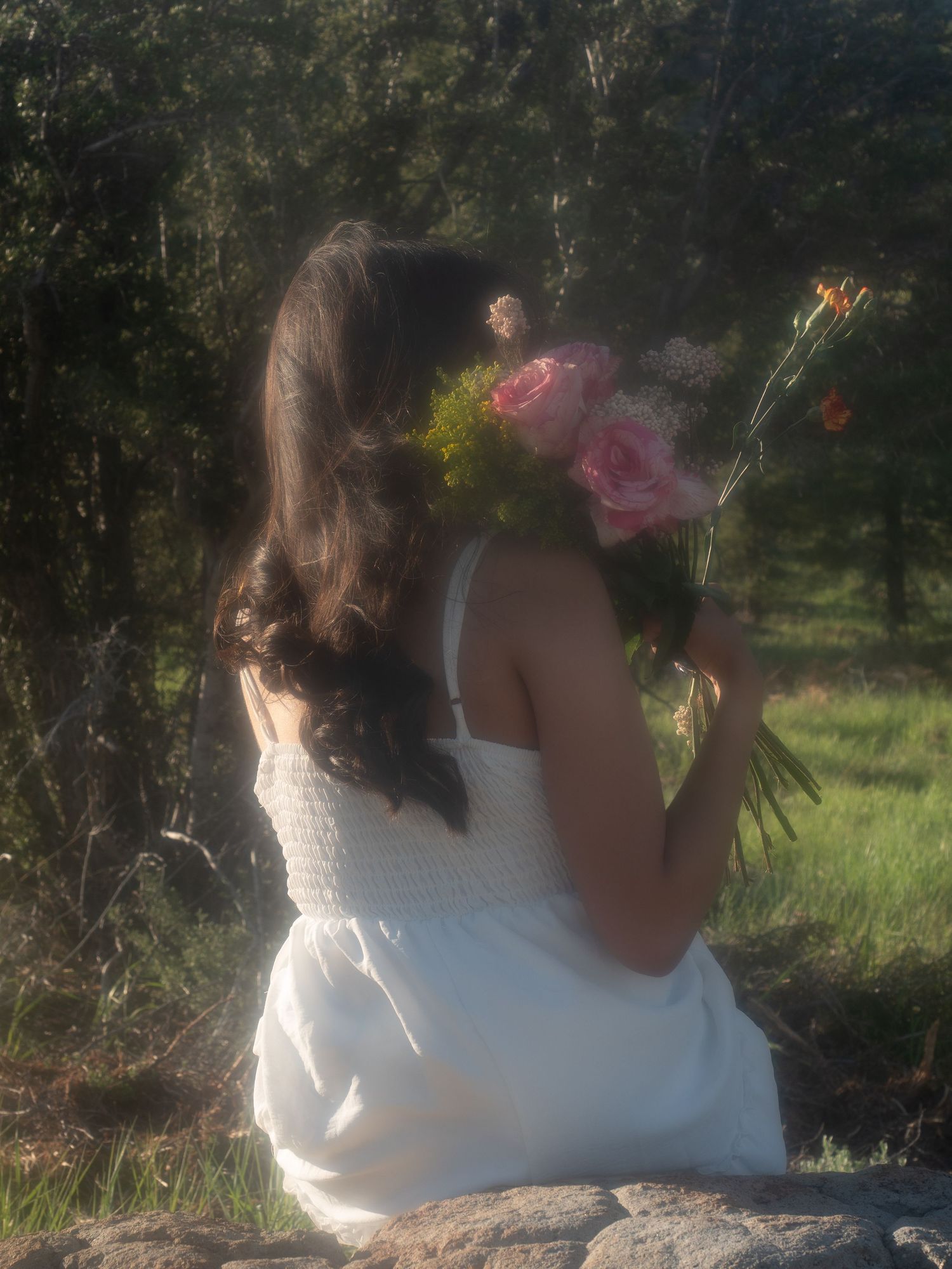 portrait woman in white dress smiling in field sitting on rock holding flowers facing away