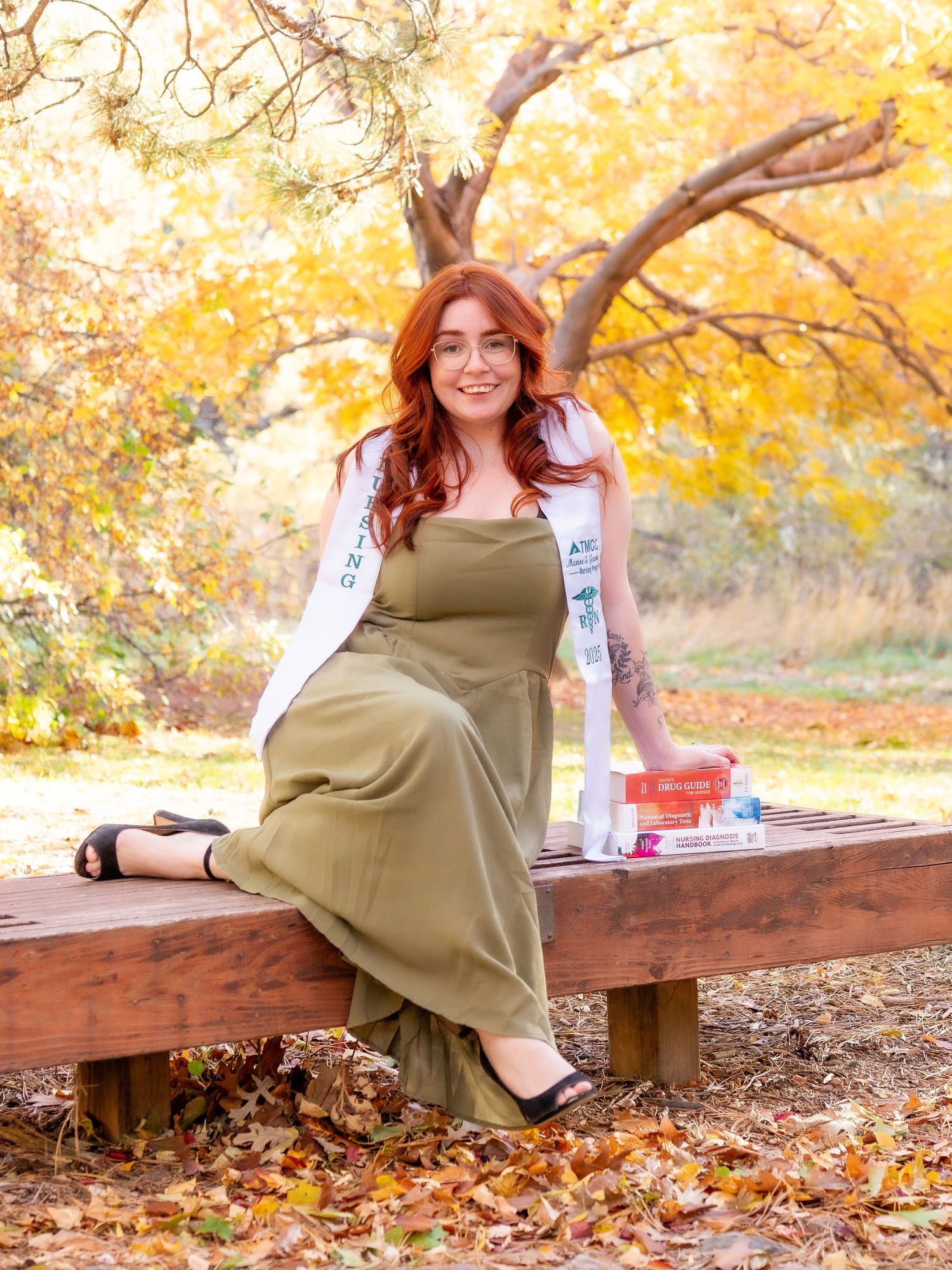 portrait graduate photo woman smiling leaning on stack of books, fall background green dress