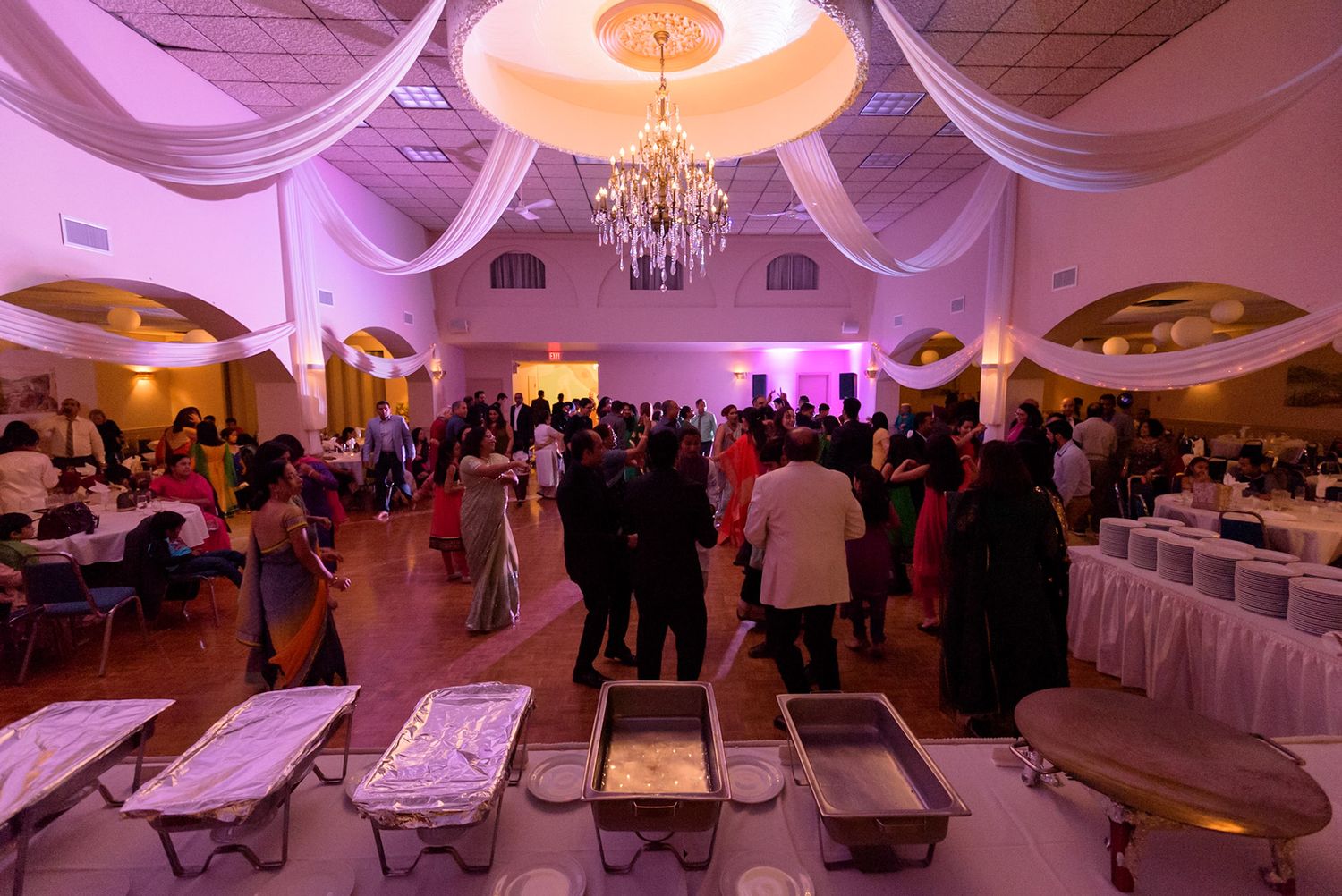 Elegant ballroom with pink uplighting, white drapes, and crystal chandeliers set up for a special event.