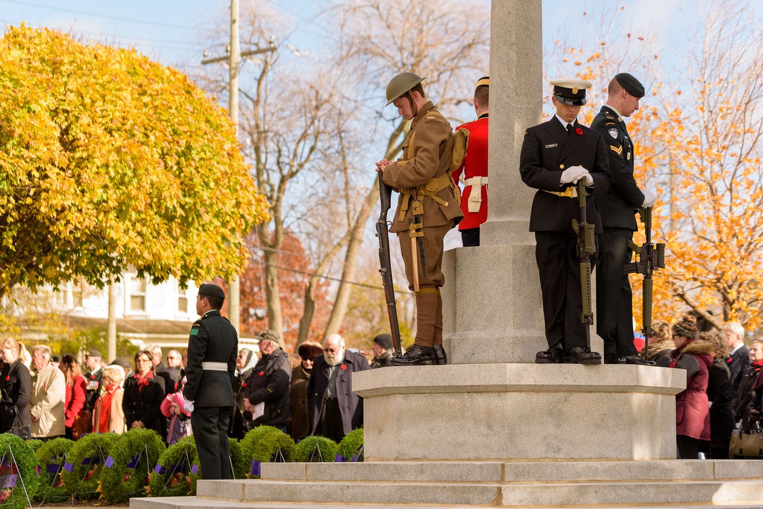 People gather around a memorial or monument during autumn with golden foliage in the background.