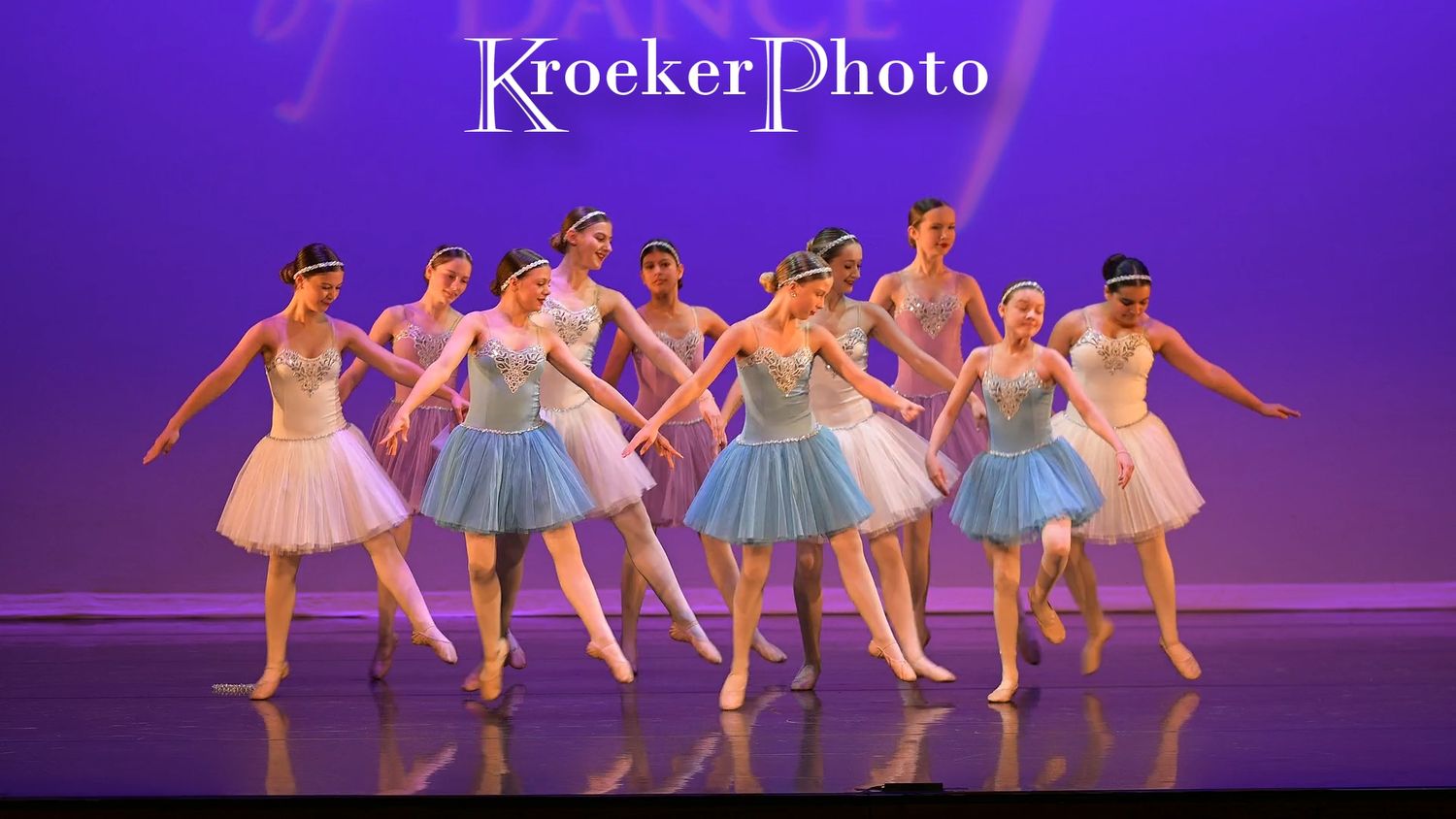 Ballet dancers in pastel tutus perform a synchronized dance routine on stage under purple lighting.