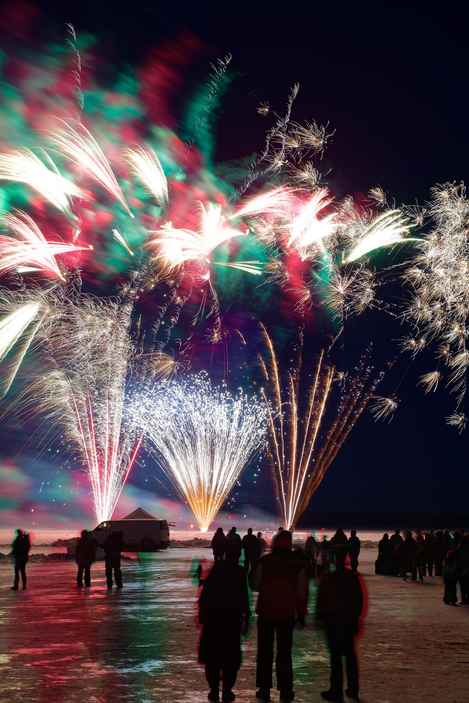 Vibrant fireworks burst over a frozen lake at night with spectators watching the display in silhouette.