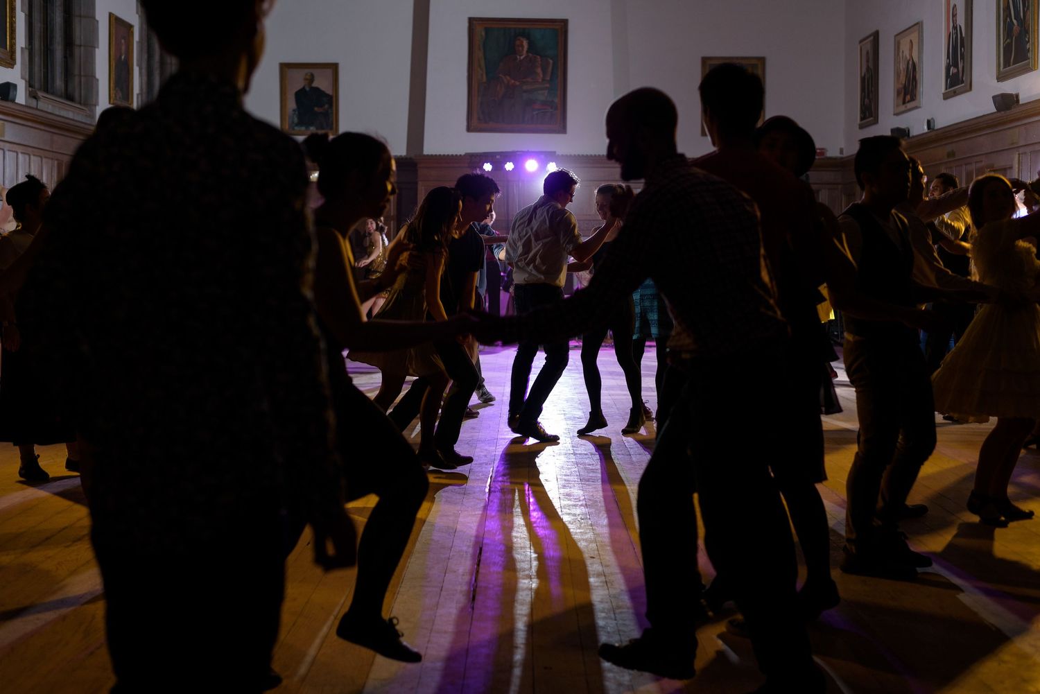 Silhouettes of dancers moving across a dimly lit ballroom with purple lighting accents.