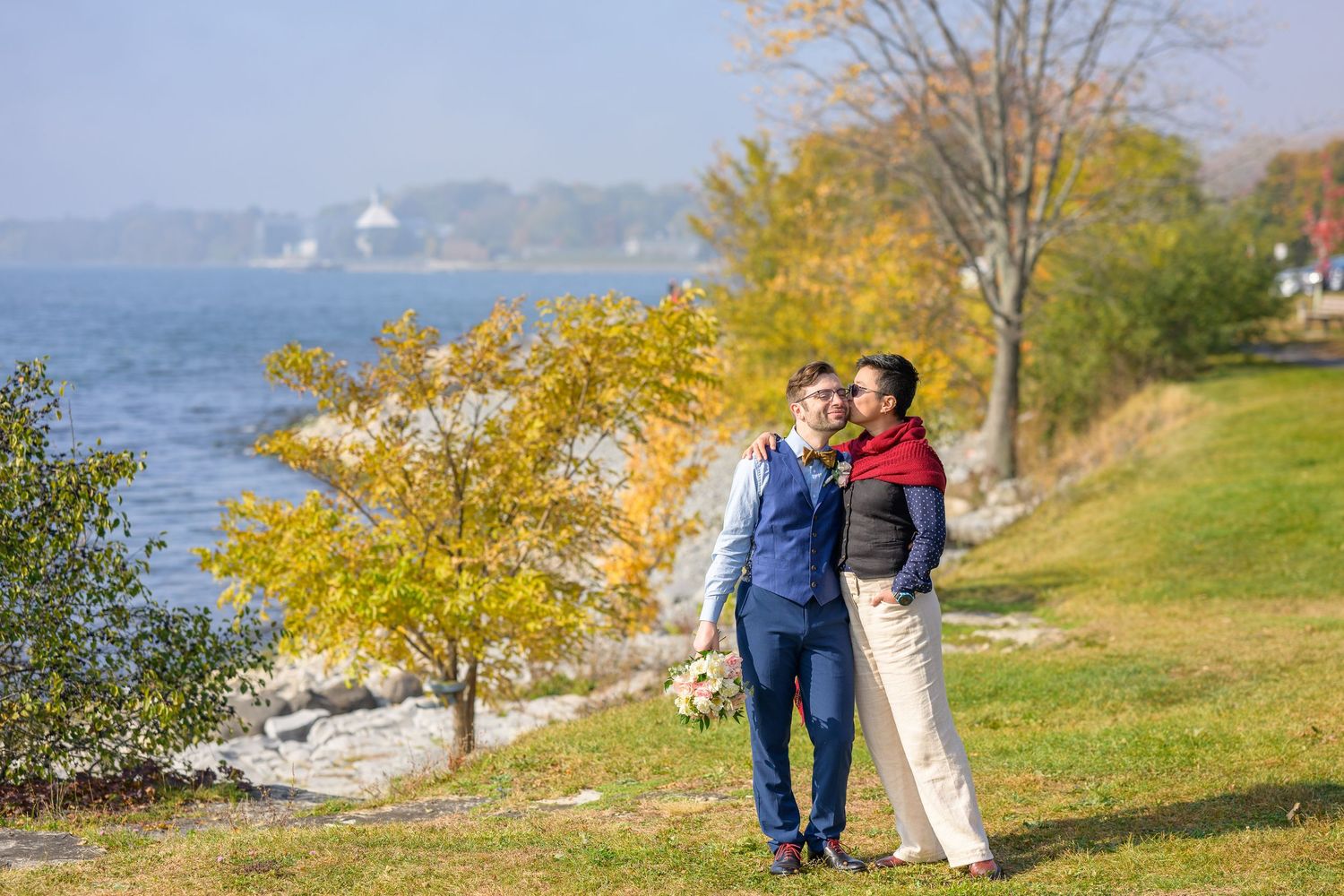 A couple walks along a scenic waterfront path with autumn trees and lake views on a sunny fall day.