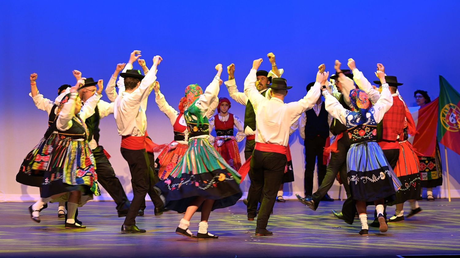 Traditional folk dancers in colourful costumes perform synchronized movements on stage under blue lighting.