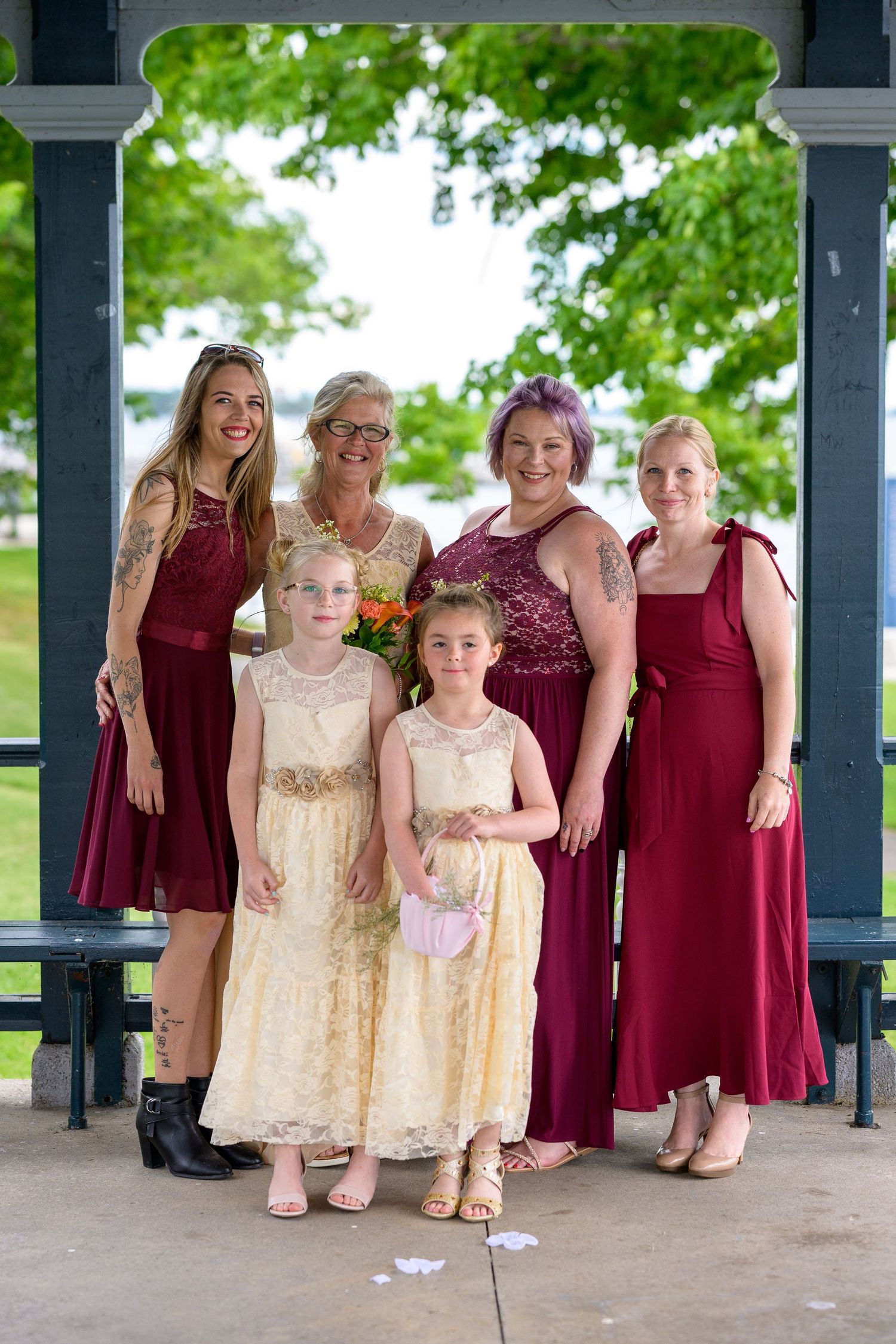 A group of bridesmaids in burgundy dresses and flower girls in cream lace dresses pose together under a gazebo.